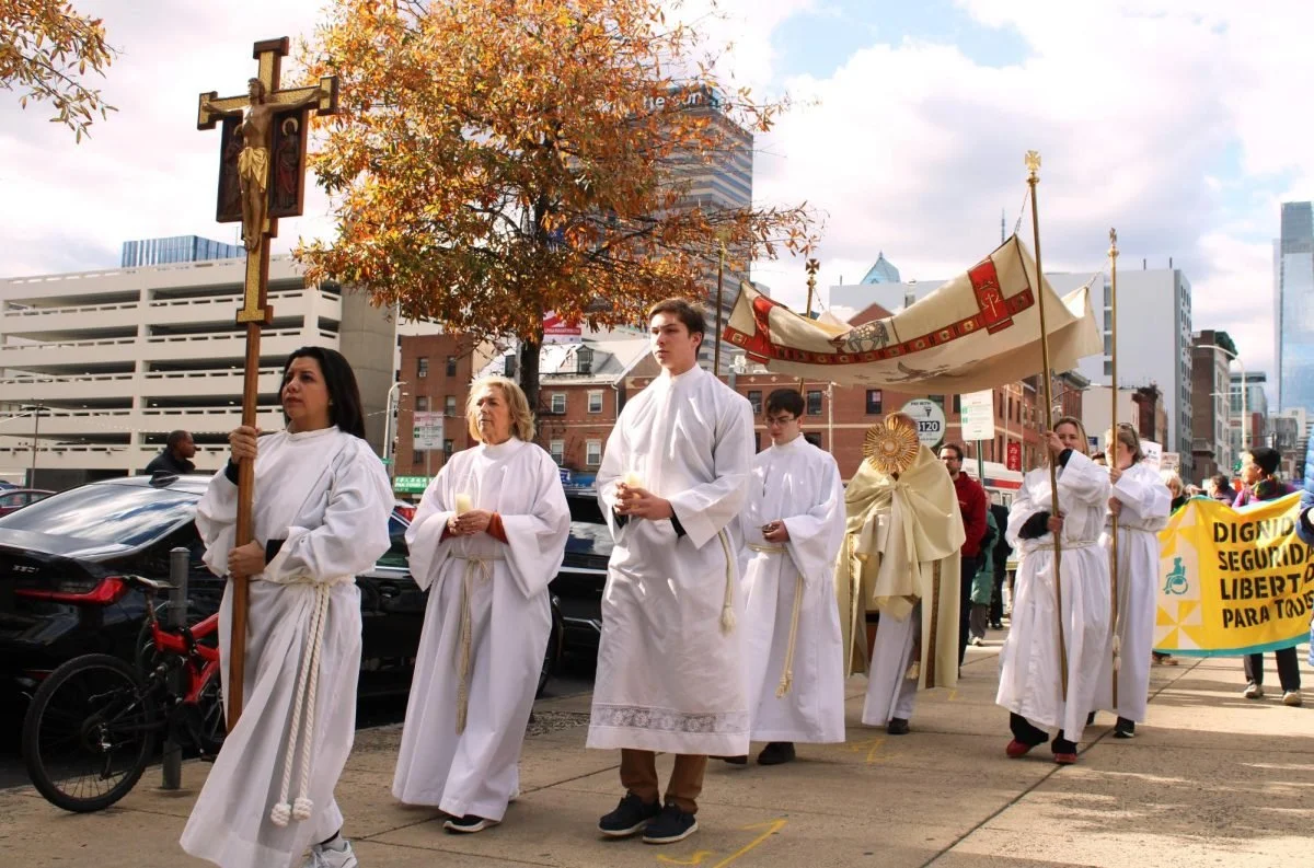People dressed in white robes participate in a religious procession on a city sidewalk, carrying religious items and banners, with a cityscape and fall foliage in the background.