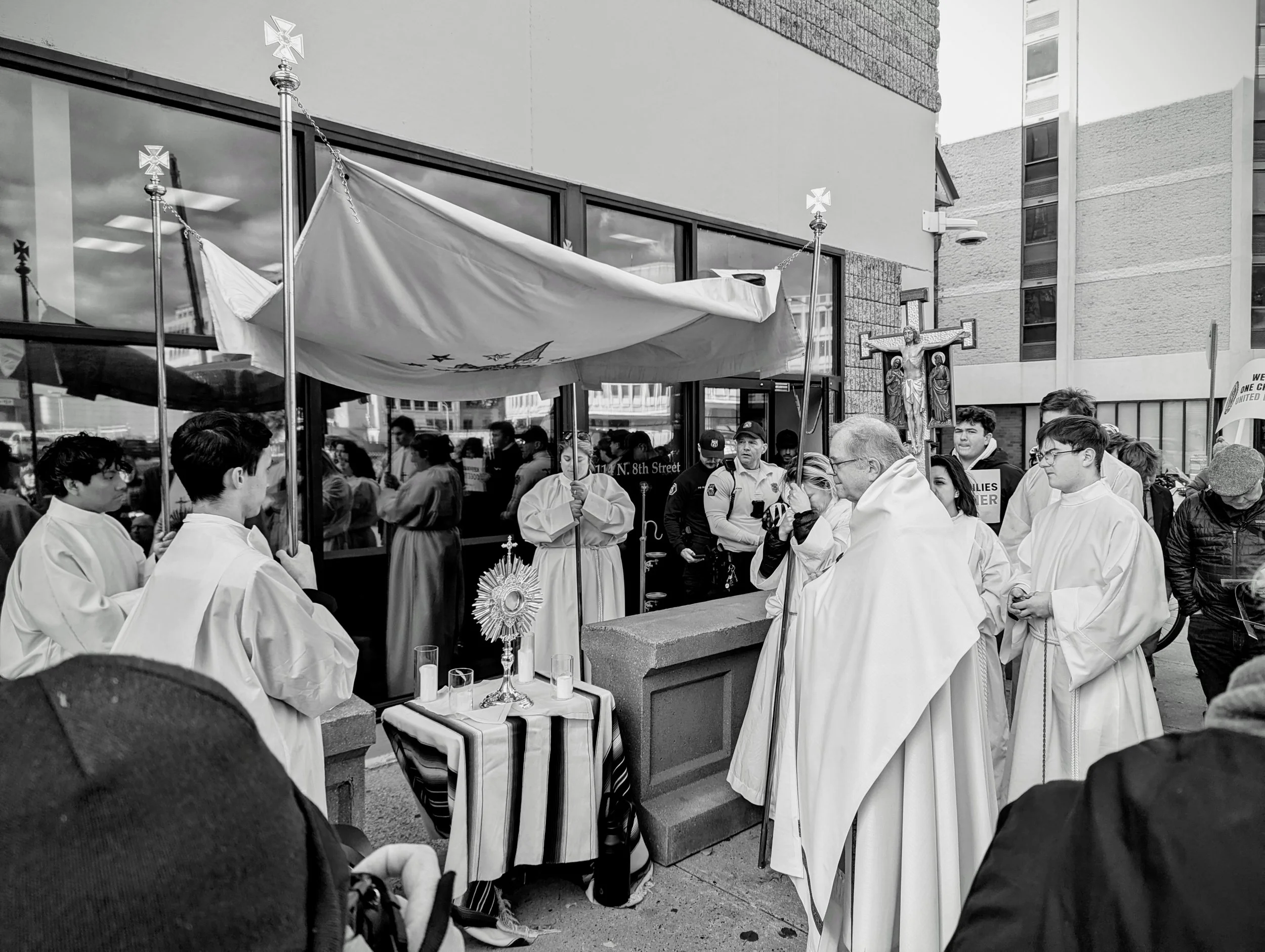 A black and white photo of a religious procession with priests and altar servers gathered around a makeshift altar, with a priest leading prayer. People are holding crosses and religious symbols, and there is a canopy overhead, in front of a modern building with large glass windows.