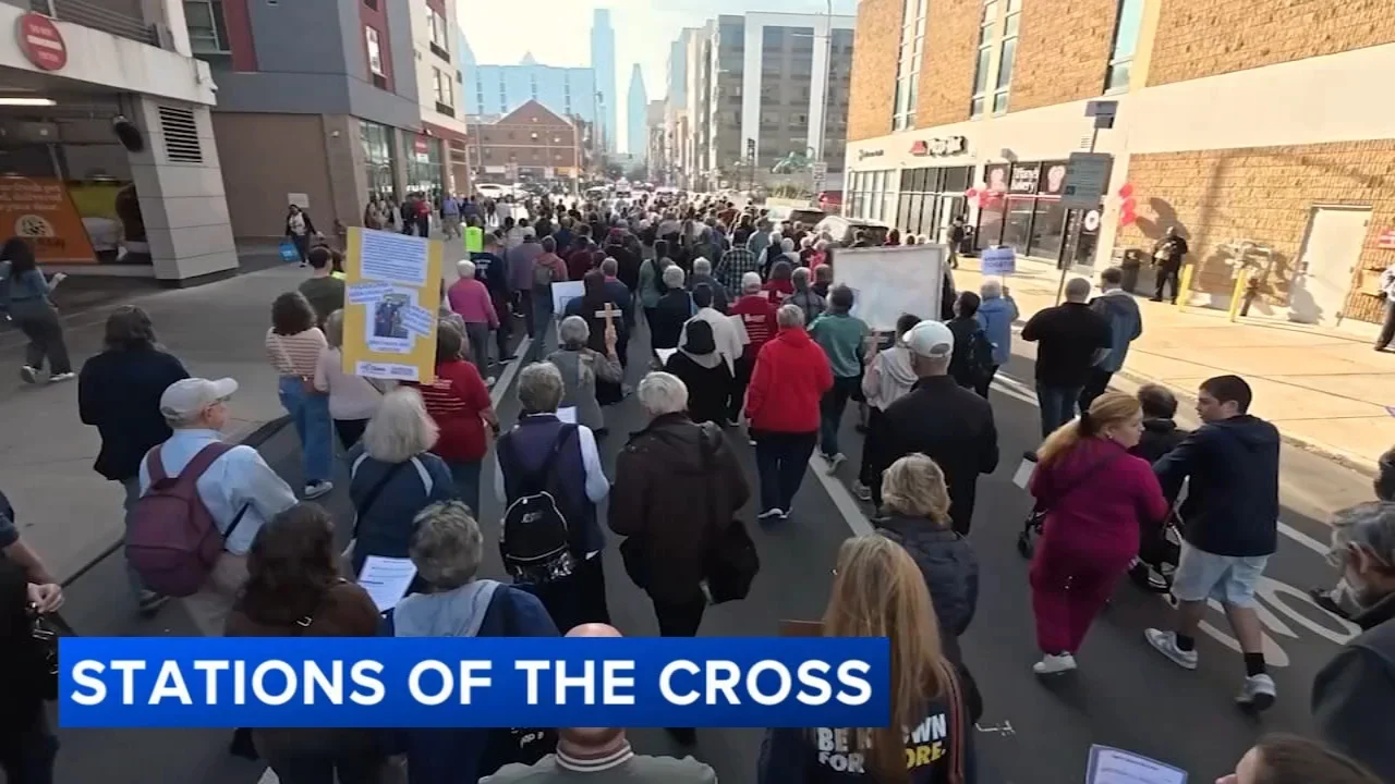 A large crowd of people participating in a march or protest on a city street during daytime. Some are holding signs, and there is a blue banner at the bottom with the text "STATIONS OF THE CROSS."