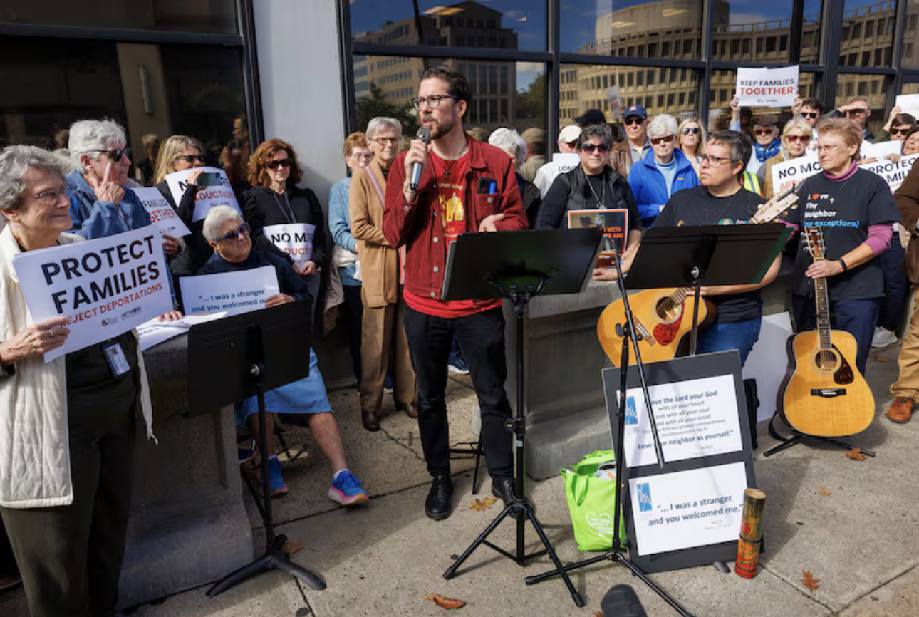 A group of people gather outside a building for a protest or rally, with signs supporting families and opposing deportations. A man in a red jacket stands at a microphone, accompanied by musicians with guitars. Others hold signs that say "PROTECT FAMILIES" and "NO MORE DEPORTATIONS."
