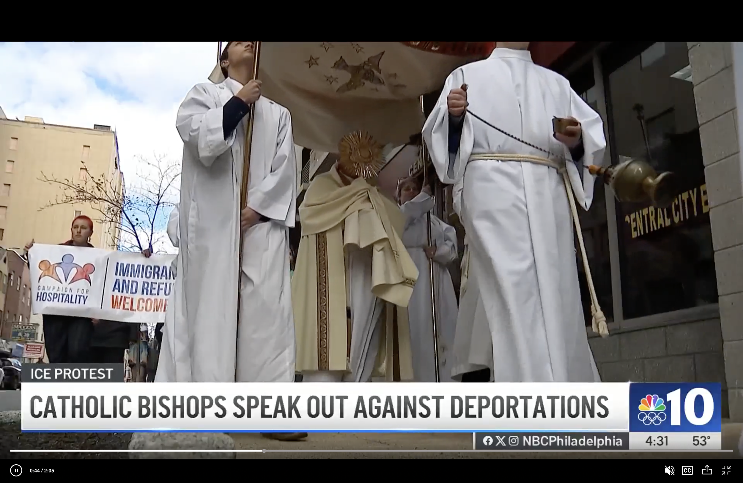 Catholic bishop dressed in religious robes holding a monstrance during a protest. Several people in white robes are nearby, and one protester in the background holds a sign that says 'Immigrants and Refugees Welcome.' The banner in the bottom-left corner reads 'ICE Protest' and the headline below states 'Catholic bishops speak out against deportations.'