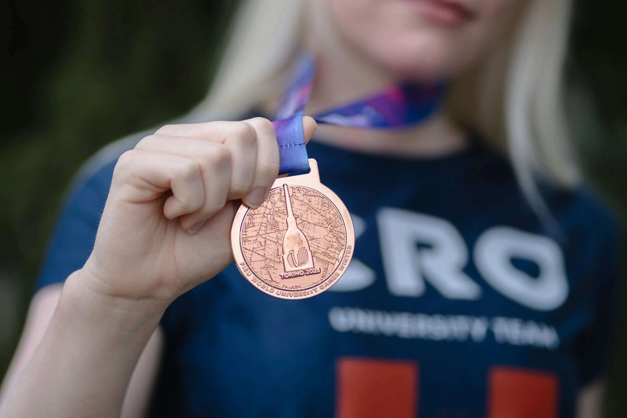 Person holding a bronze medal with a blue ribbon, showing a fork and the words 'Torino 2023' and 'PSU World University Games Wu Jie'.