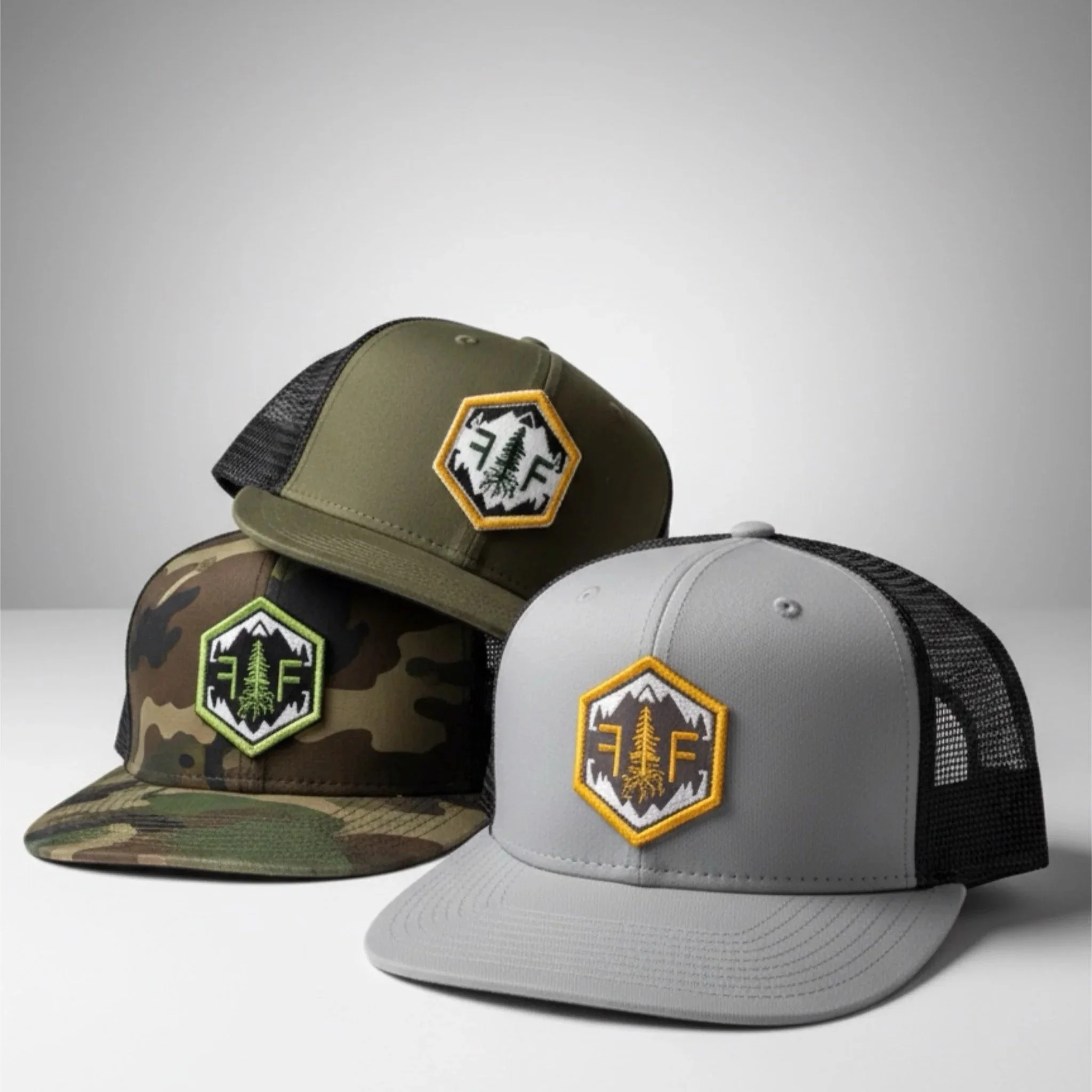 Three trucker hats with outdoor-themed patches, one in camouflage, one in olive green, and one in light gray.