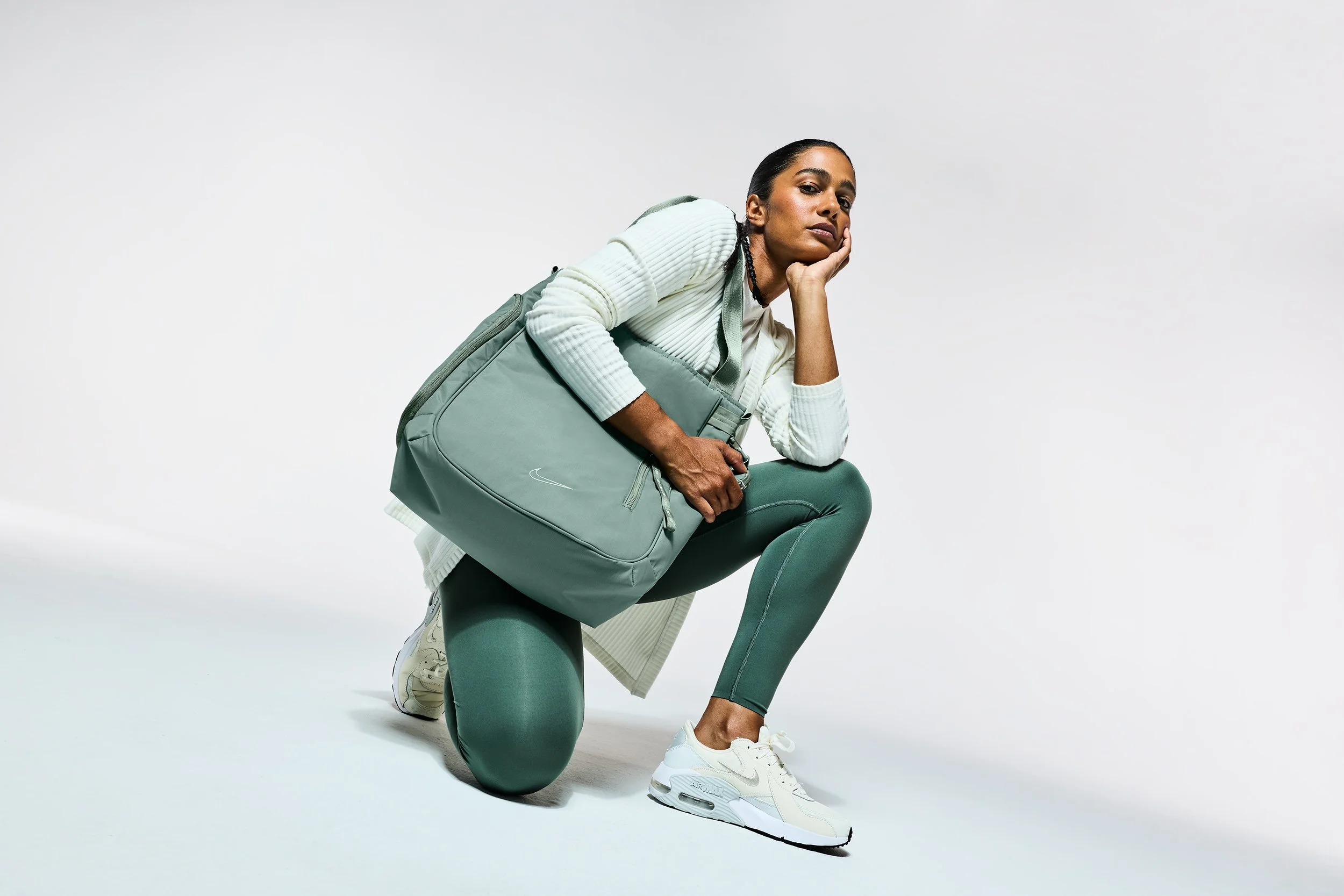 A woman in athletic wear kneeling on one knee with a large gray gym bag, wearing white sneakers, against a plain white background.