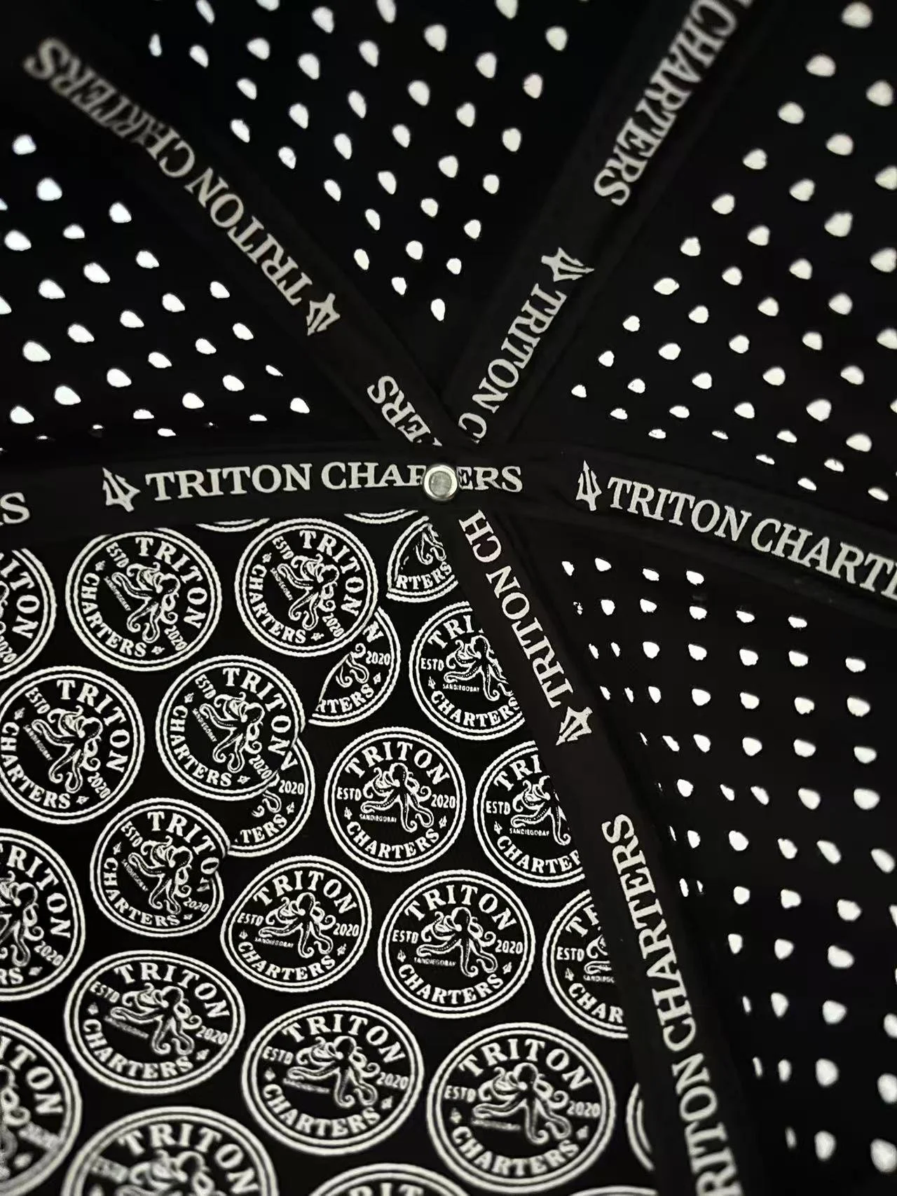 Close-up view of black fabric with white polka dots and the words 'A TRITON CHARTERS' printed on the seams, along with a pattern of circular logos reading 'TRITON CHARTERS ESTD 2020' and a trident logo.