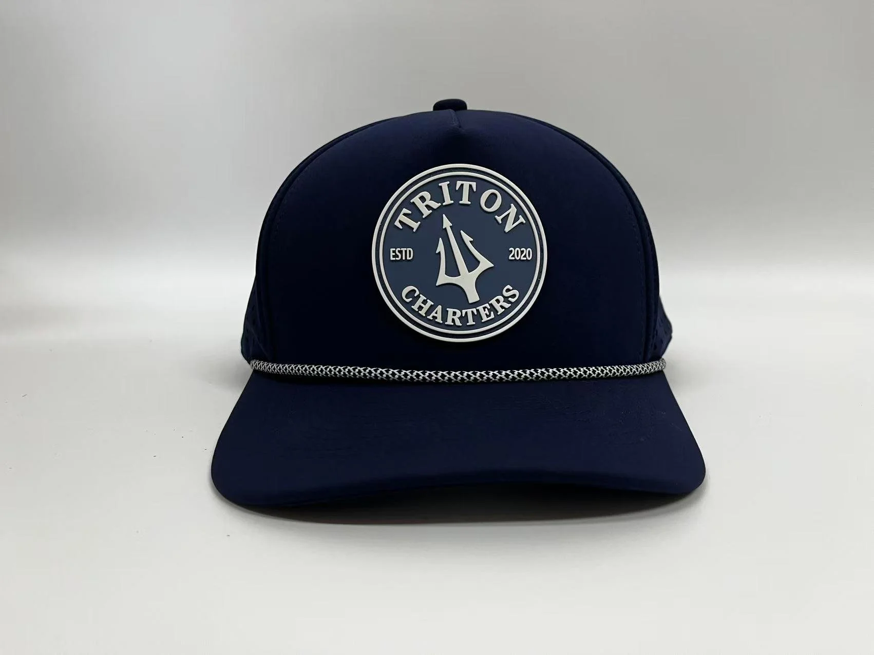Navy blue baseball cap with a round patch on the front that reads 'Triton Charters' with a trident symbol, ESTD 2020, and a decorative braid along the brim.