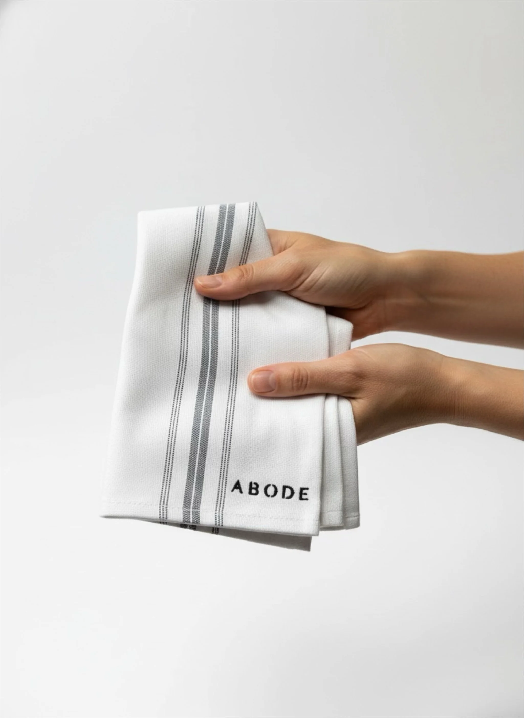 A pair of hands holding a folded white kitchen towel with black and gray stripes and the brand name 'ABODE' embroidered in black in the lower corner.