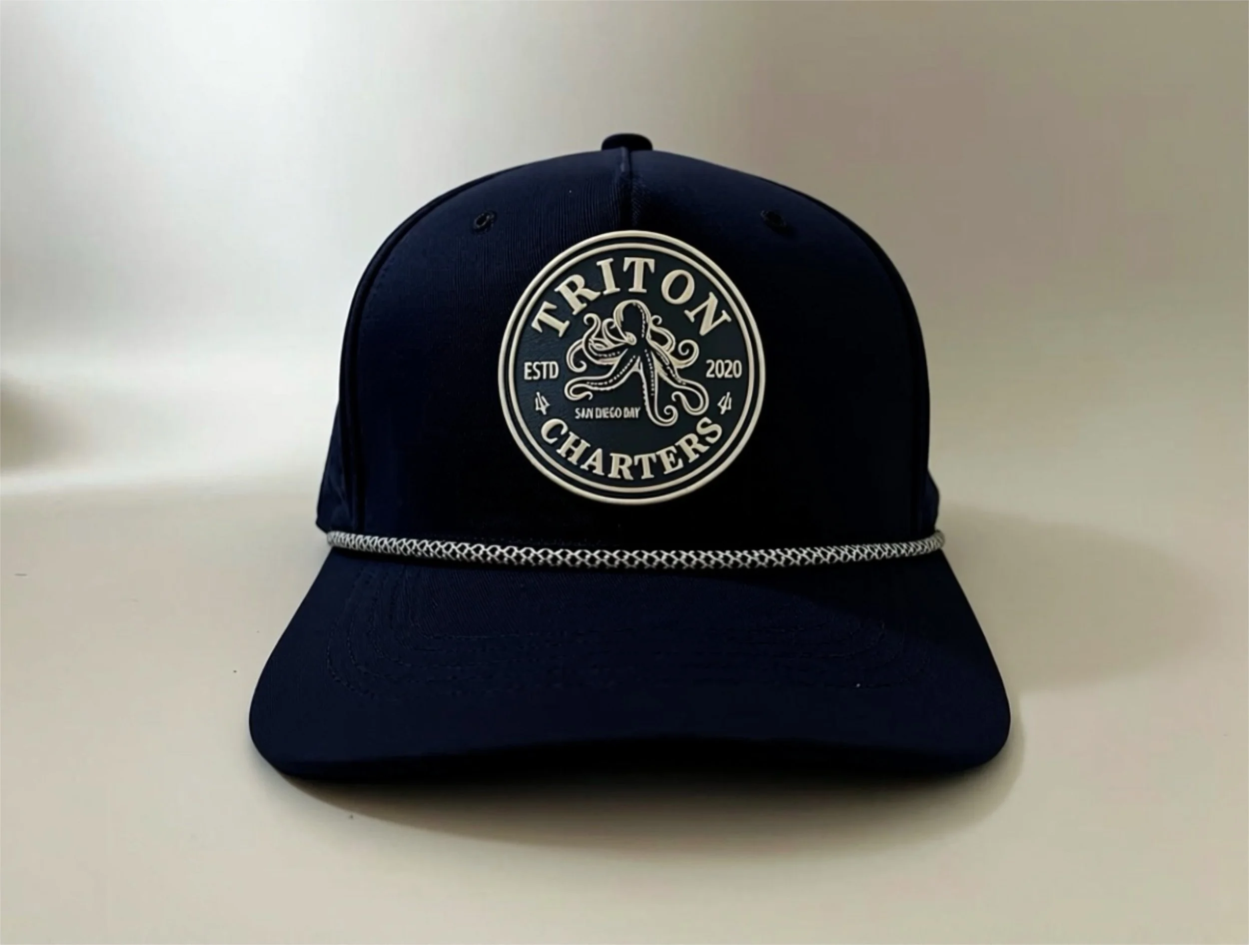 A navy blue baseball cap with a circular logo patch on the front, featuring an octopus, the text "Triton Charters," "Est  2020," and "San Diego Bay," and a decorative brim detail.