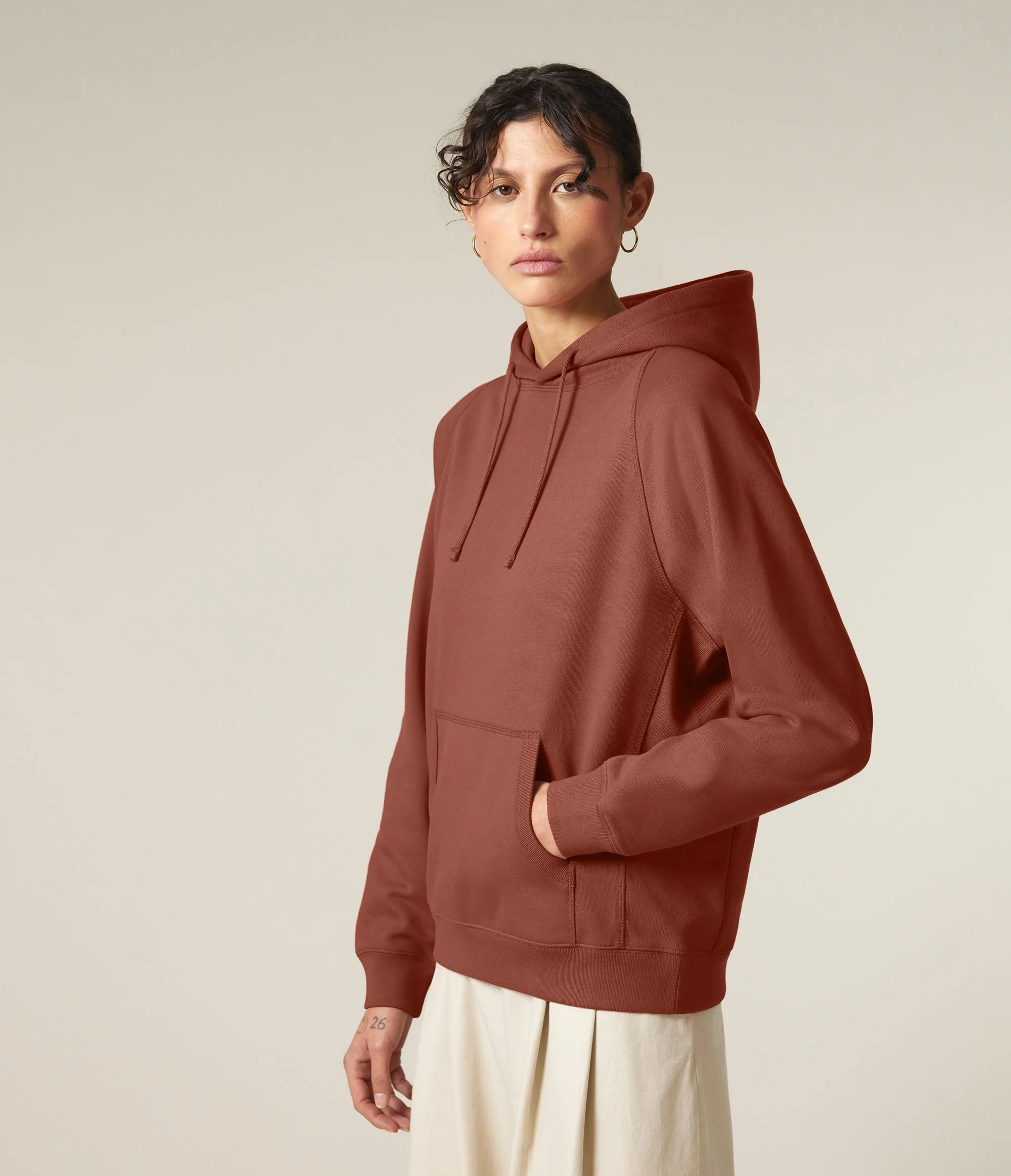 A woman wearing a rust-colored hoodie with a front pocket and drawstrings, paired with white pants, standing against a neutral background.