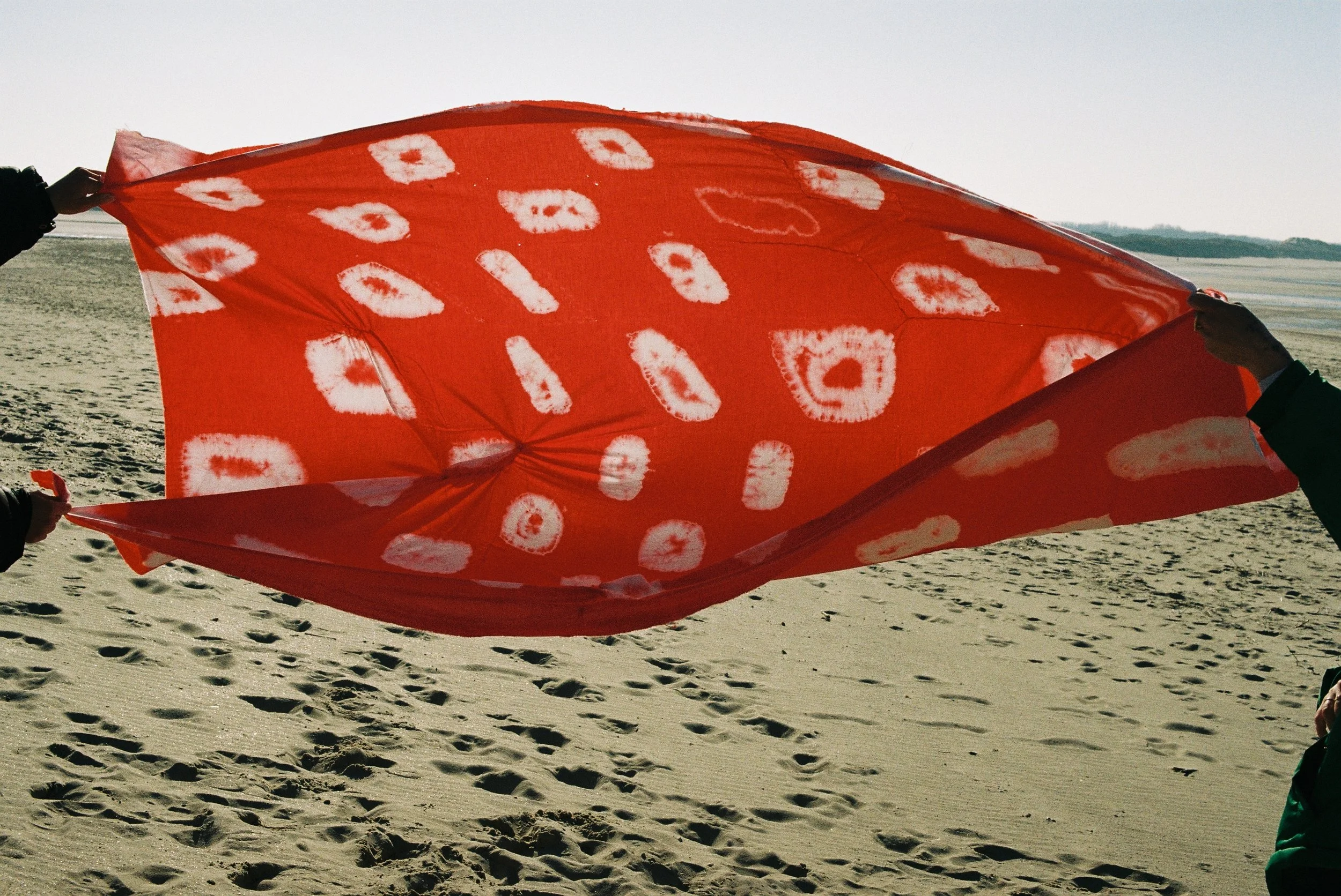 Deux personnes tenant un cerf-volant rouge avec des motifs blancs dans un paysage désertique ou plage.