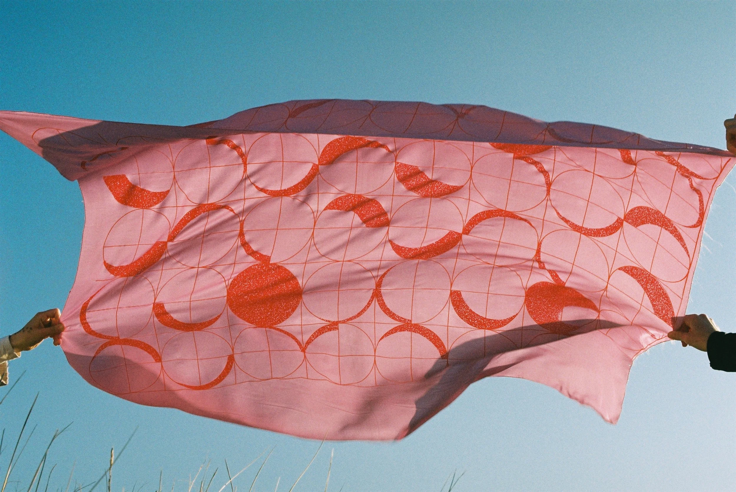 Two people are holding a piece of fabric made using the serigraphy technique, featuring red circular patterns — moon cycles ; the fabric is fluttering in the breeze on a North Sea beach.