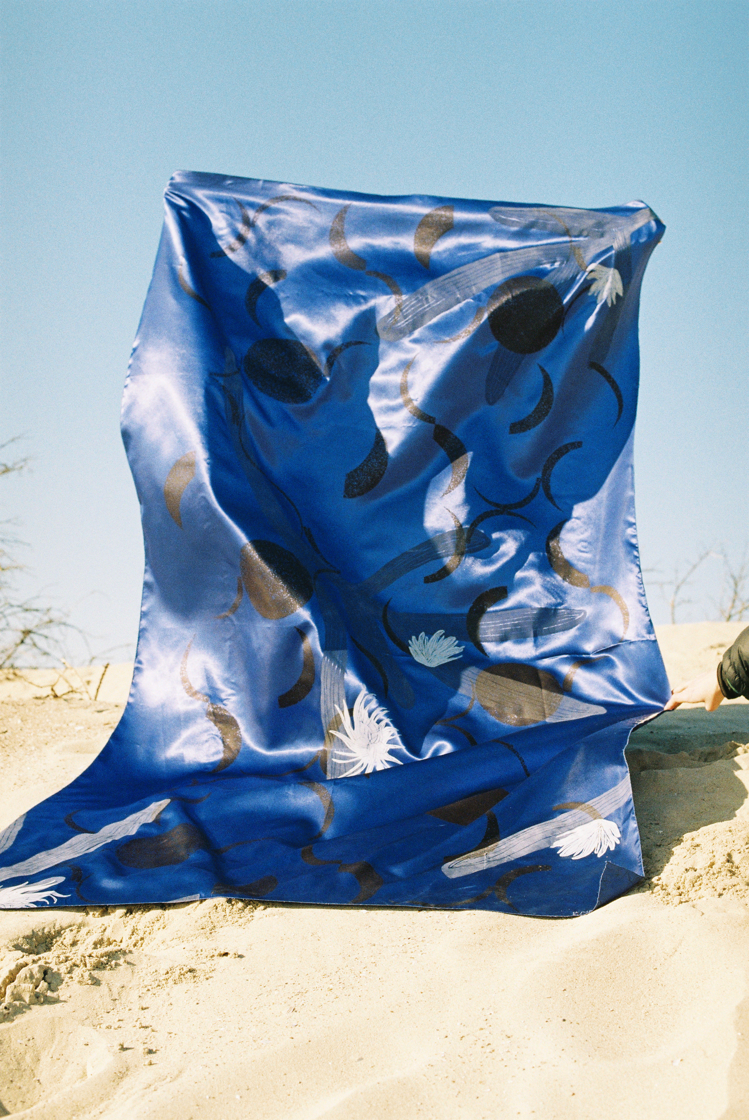 A large, satin-blue screen-printed fabric featuring floral patterns and abstract shapes flutters in the breeze on a sandy beach, beneath a clear sky.