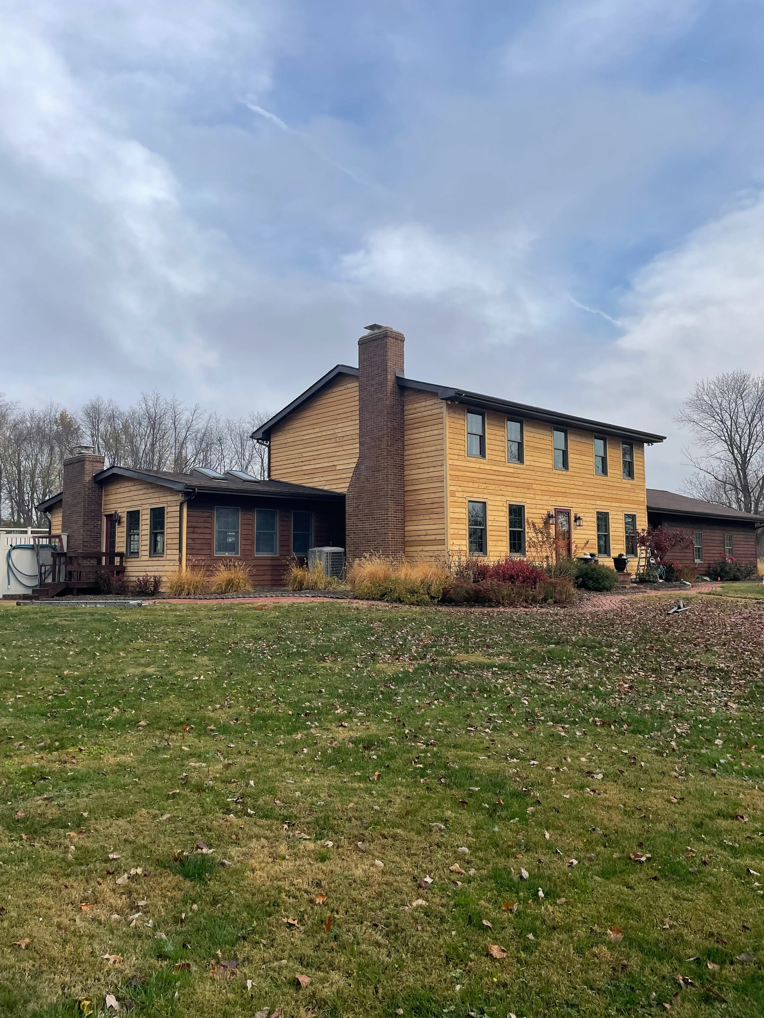 A two-story house with a large brick chimney, a mix of wood siding, and a lawn with fallen leaves, under a partly cloudy sky.