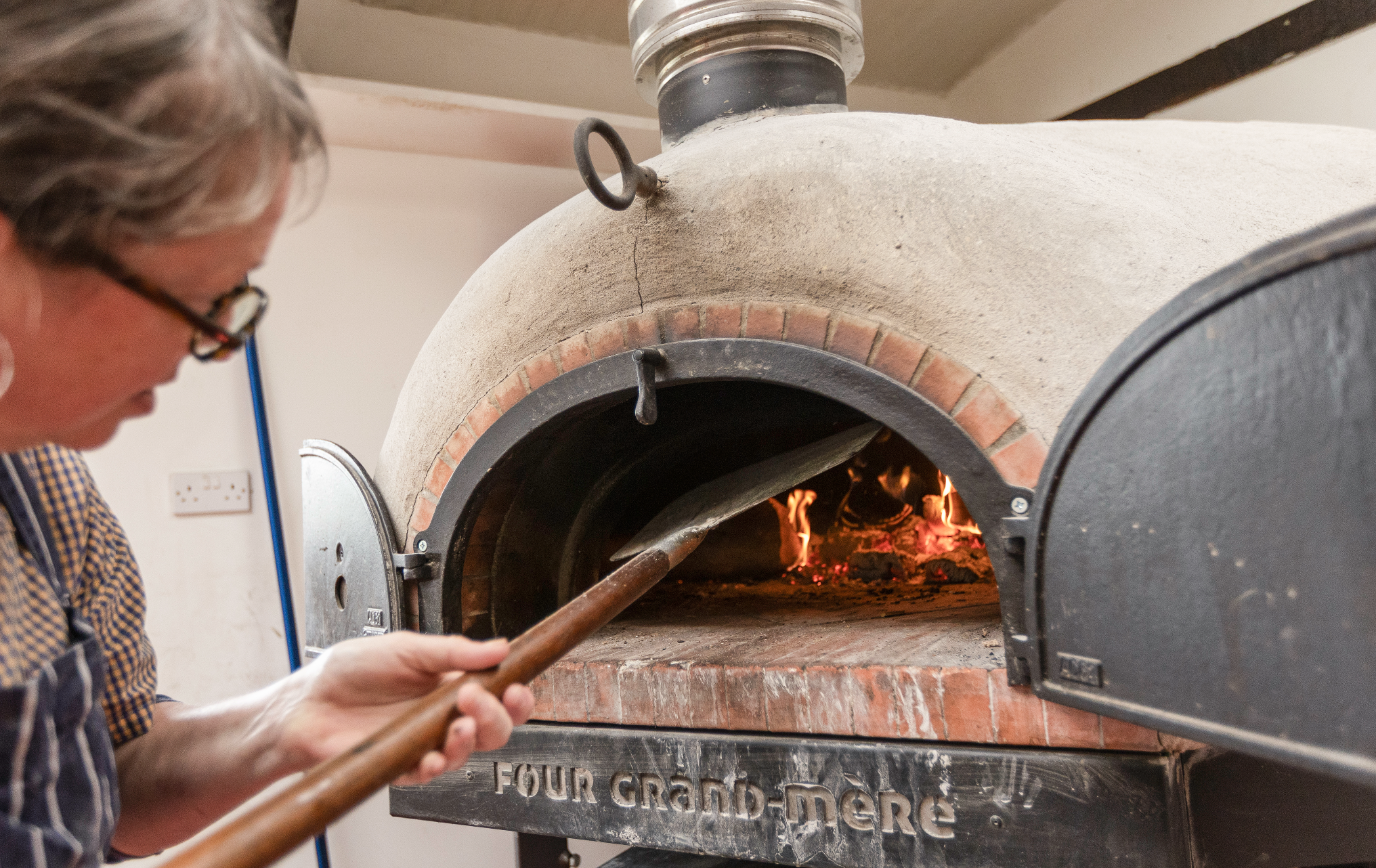 A person wearing glasses and a checkered shirt making pizza dough in front of a brick oven with a fire inside, with the oven's name, 'Four Grand-Mère', visible on the front.