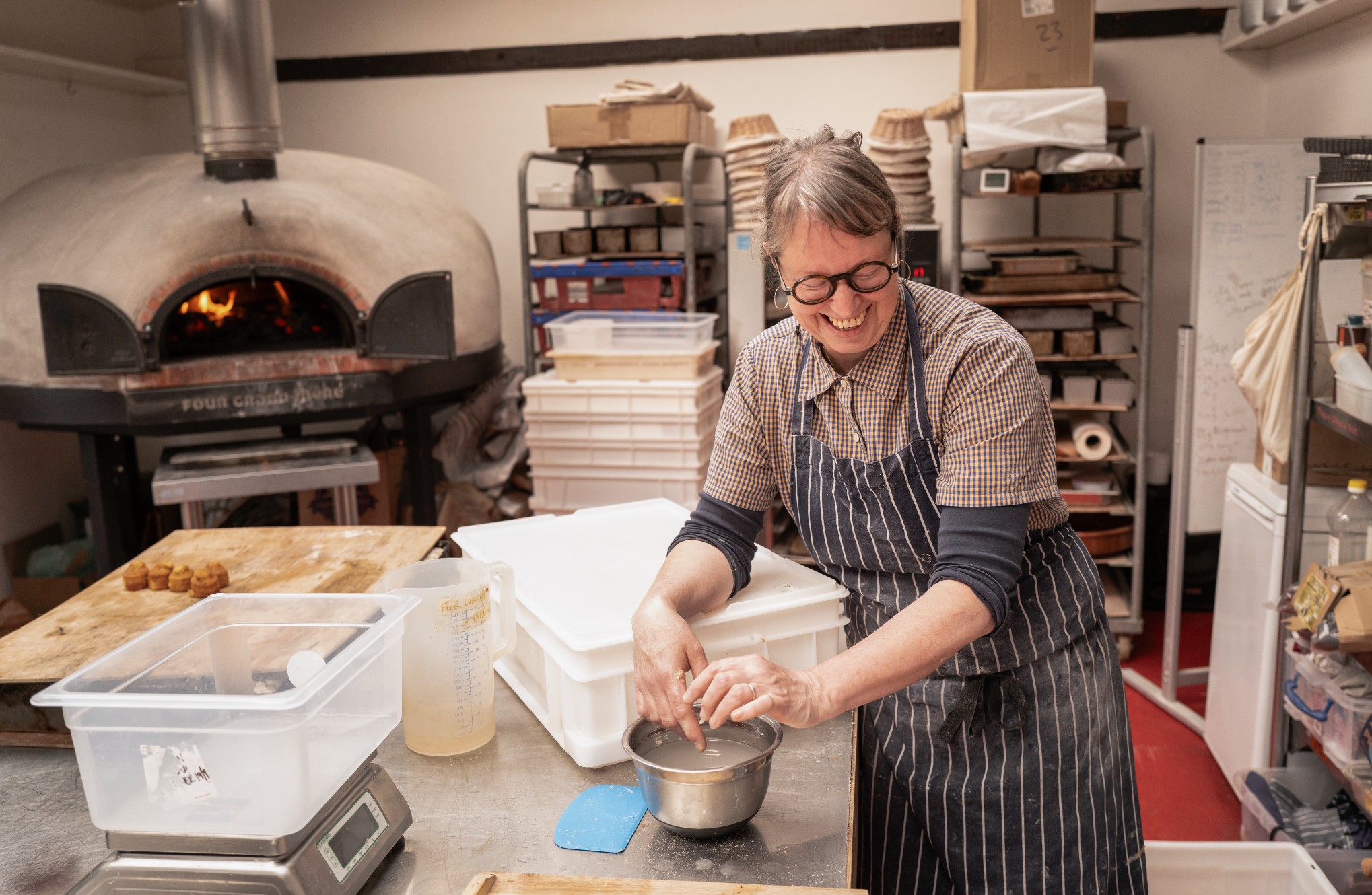 A woman with short gray hair and glasses, wearing a checkered shirt and an apron, is baking in a kitchen. She is smiling and working with dough or clay at a table with various containers and a scale. Behind her are shelves with baking trays, boxes, and kitchen supplies, and a wood-fired oven with an open door showing a fire inside.