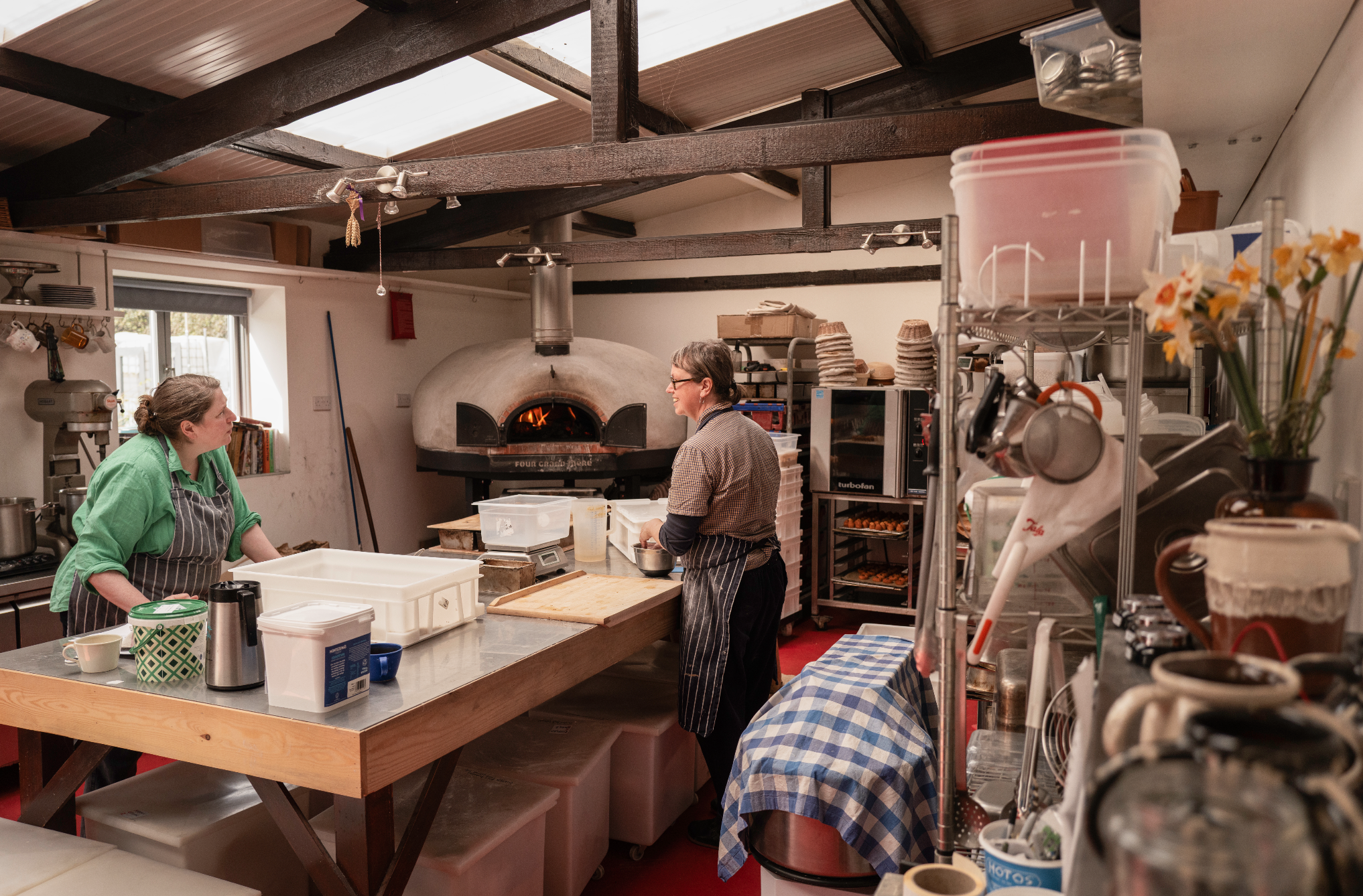 Two women working in a busy kitchen with a large pizza oven in the background, surrounded by shelves with kitchen supplies and tools.