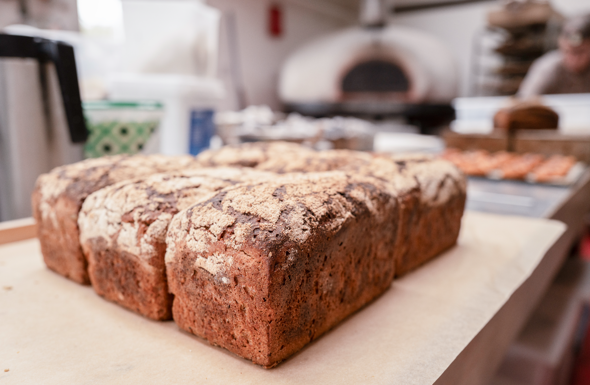 Close-up of a freshly baked loaf of bread with a cracked crust, sitting on a white surface in a bakery kitchen.