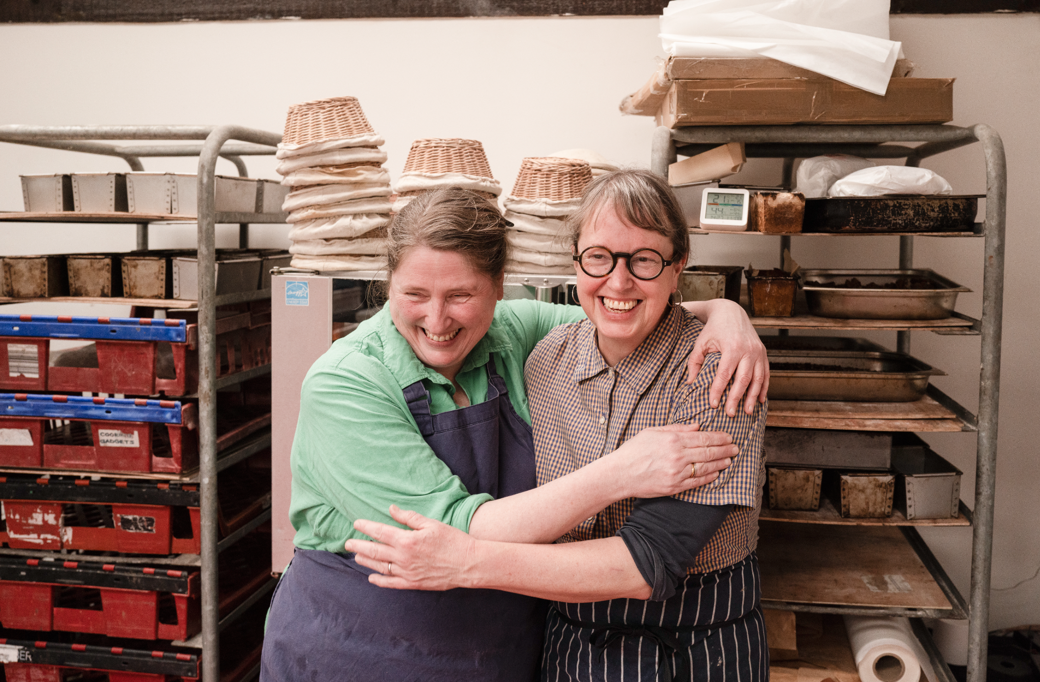 Two people, a woman with glasses and a woman with a green shirt, hugging and smiling in a bakery or kitchen setting with shelves of baked goods and trays in the background.