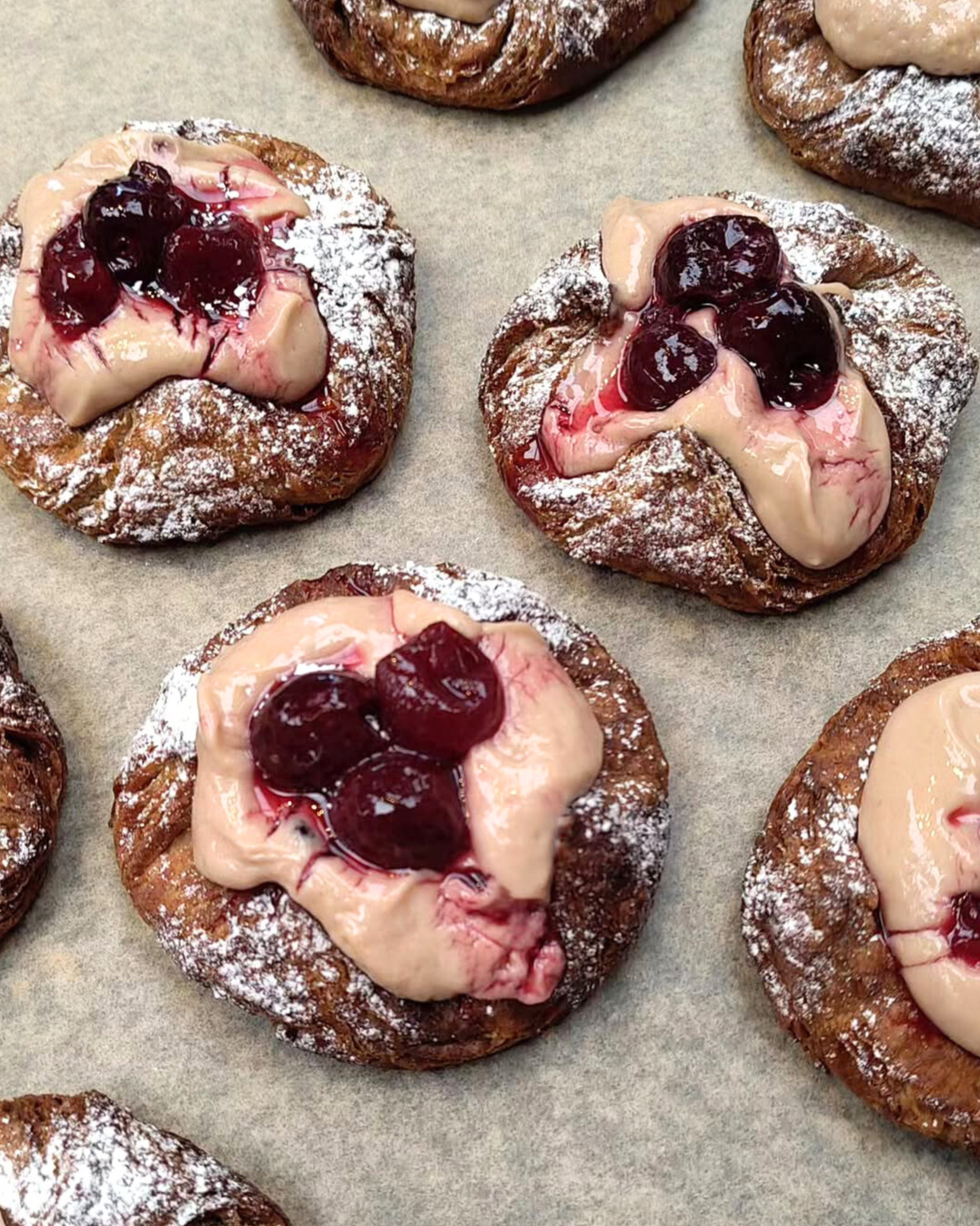 Close-up of freshly baked Danish pastries topped with cherries, cream, and dusted with powdered sugar on a baking sheet.