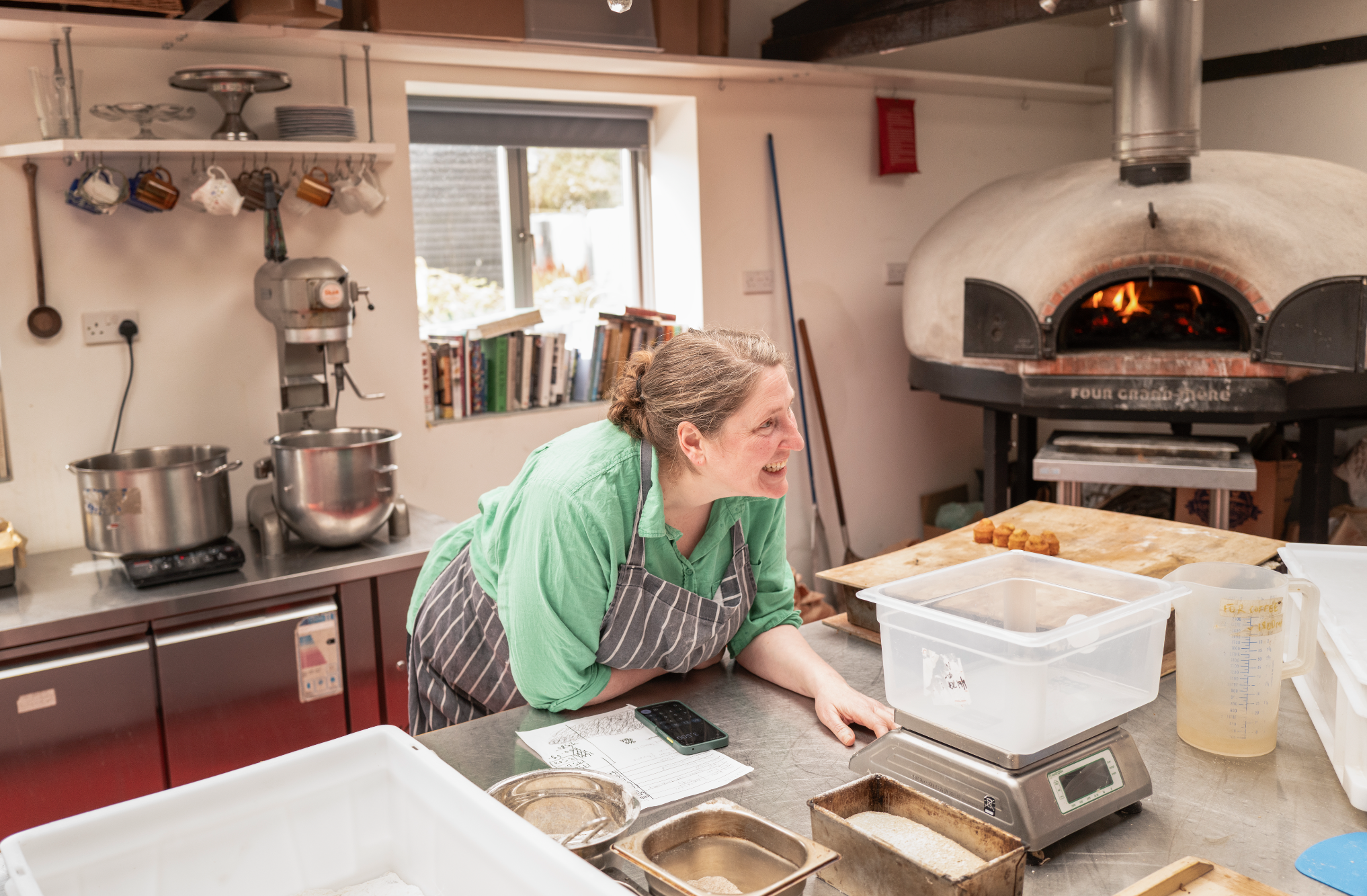 Woman smiling in a kitchen with a pizza oven, kitchen utensils, books, and various kitchen items on countertops and shelves.