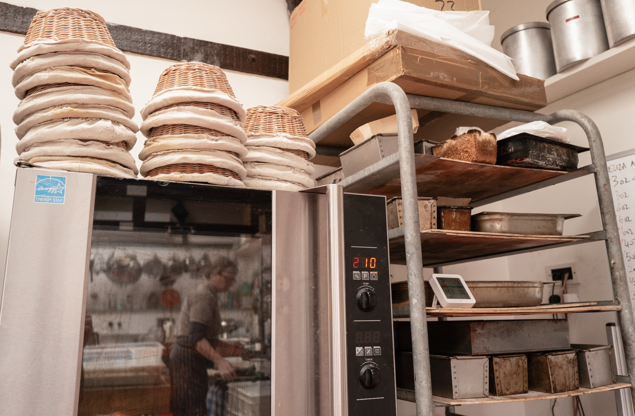 Stacks of uncooked pizza dough on a metal rack in a kitchen. A microwave oven with a digital display showing 210 is visible in the foreground. Metal pans and boxes are on the shelves above and below the rack.