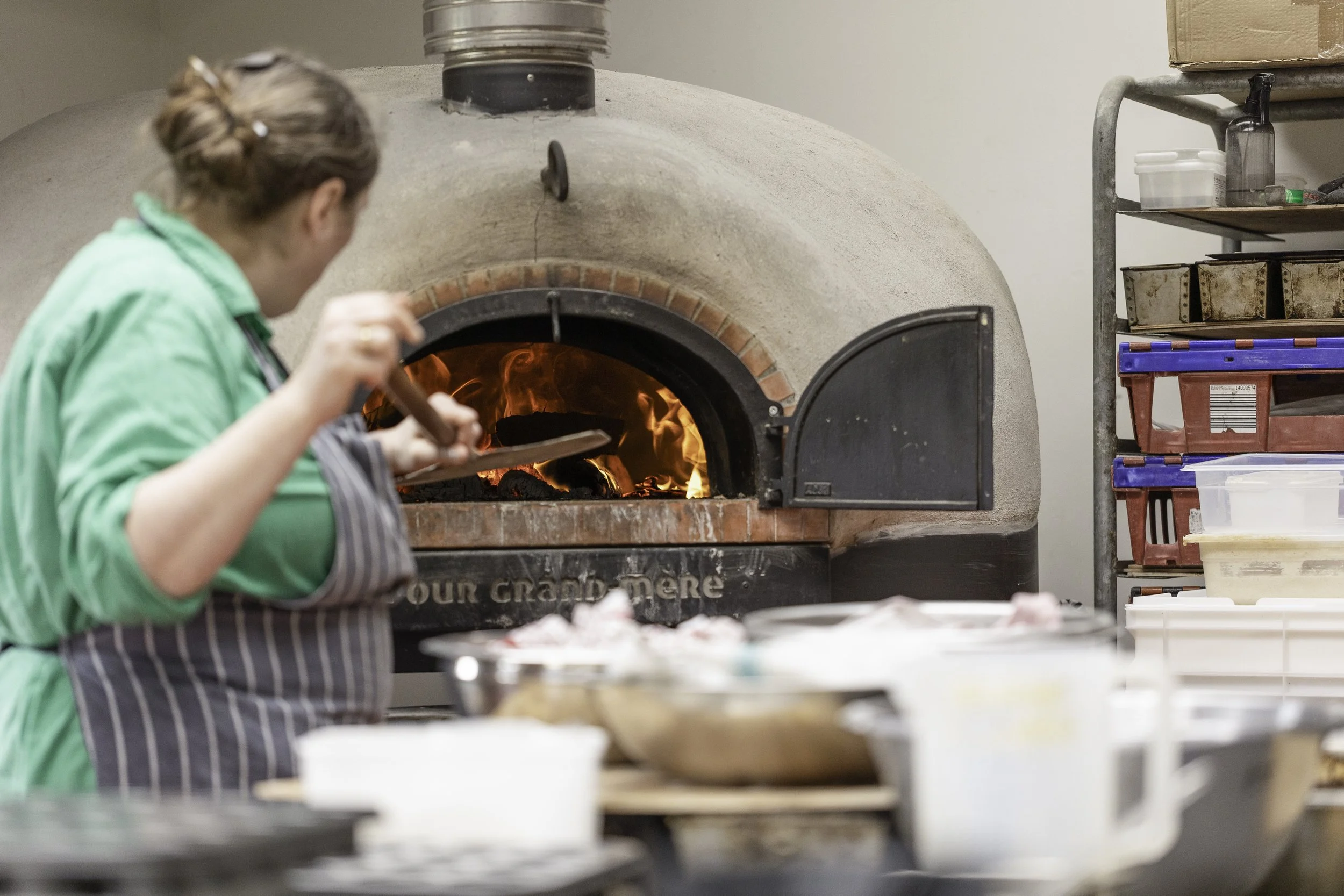 A person in a green jacket and apron working in a pizzeria oven. The oven has a stone exterior and an open door with visible fire inside. There are shelves with boxes and containers to the side.