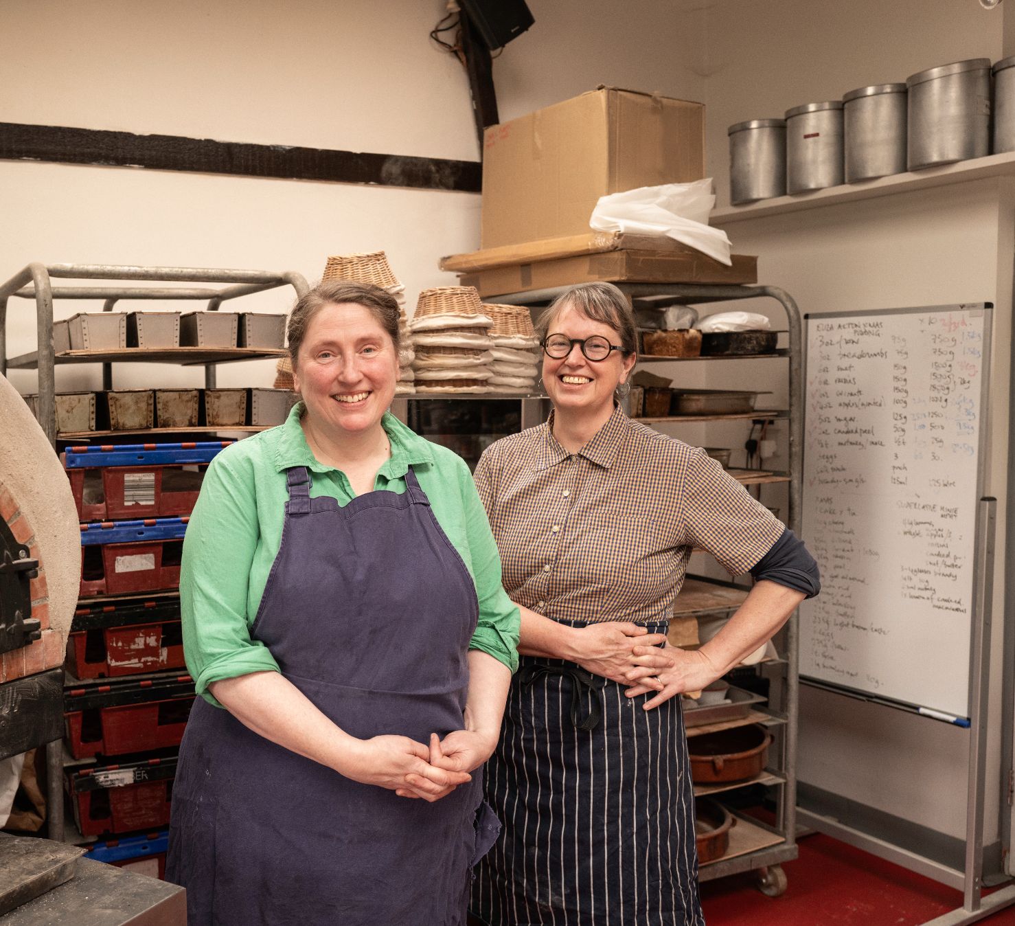 Two women standing in a bakery kitchen, smiling. The woman on the left wears a green shirt and purple apron, and the woman on the right wears glasses, a checkered blouse, and striped apron. Behind them are bakery racks filled with baked goods and a whiteboard with handwritten notes.