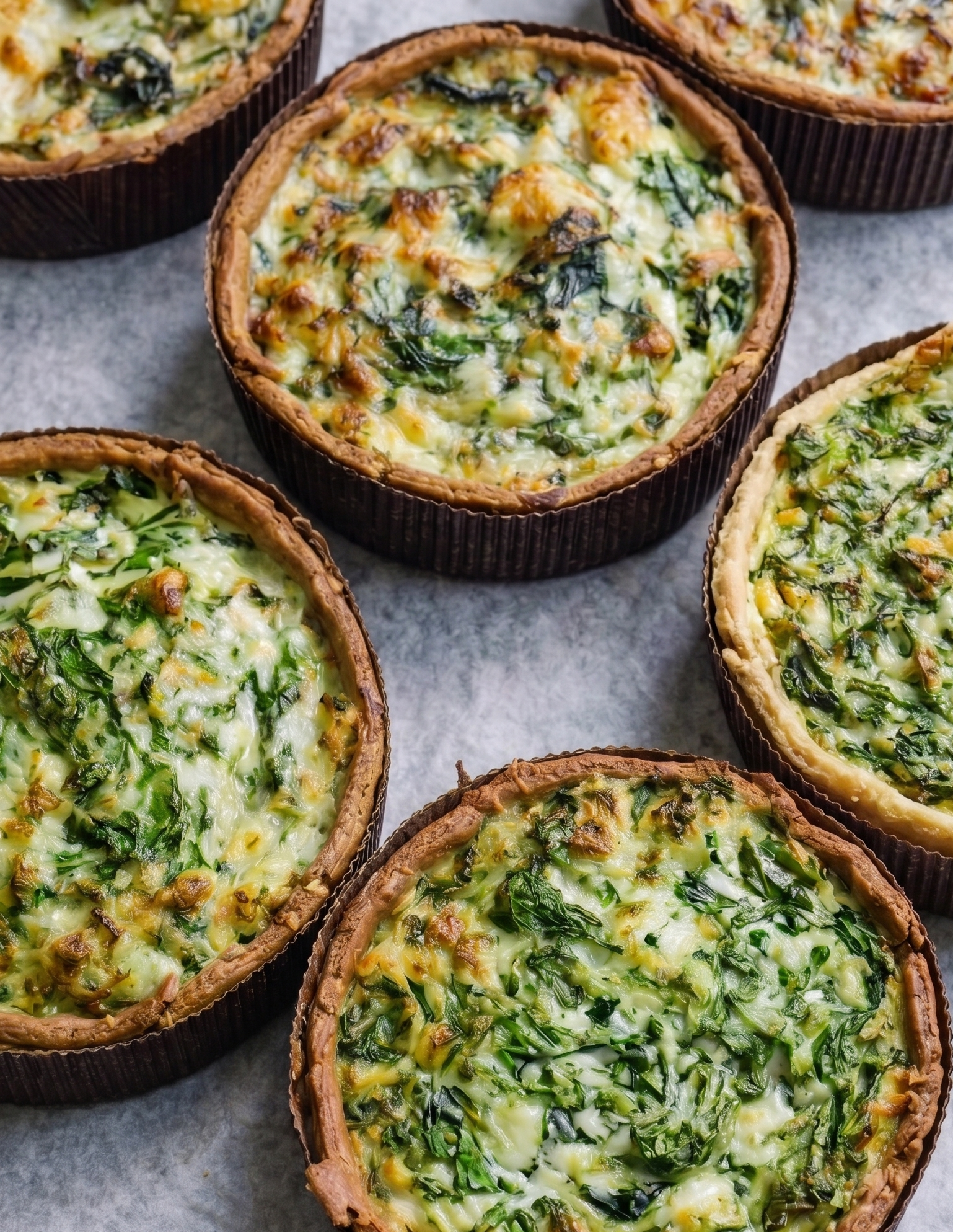 Close-up of several individual vegetable quiches with golden crusts, filled with spinach, cheese, and chopped vegetables on parchment paper.