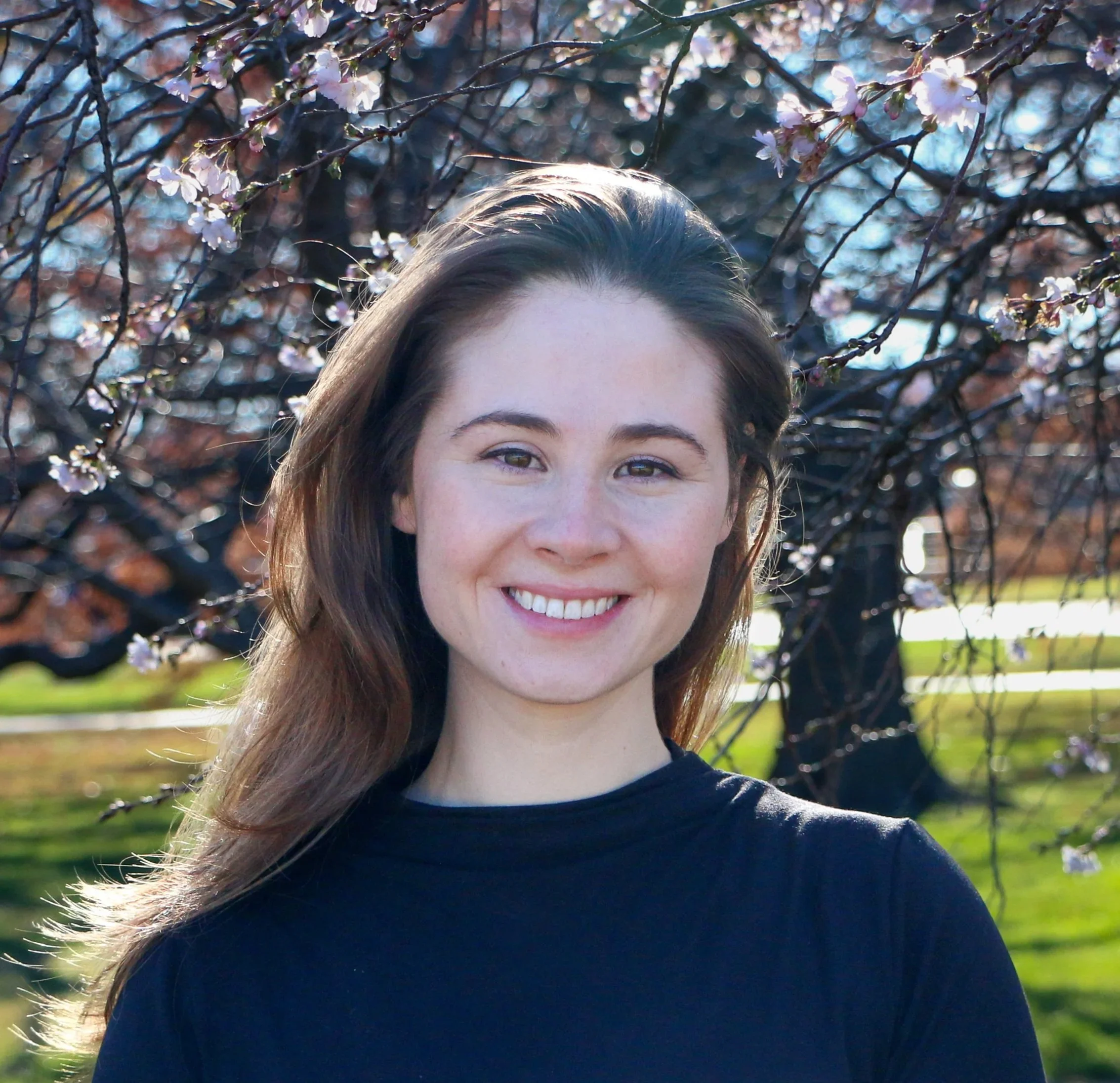 A woman with long brown hair smiling outdoors in front of blossoming tree branches on a sunny day.