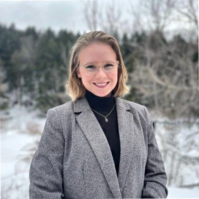 A young woman with blonde hair wearing glasses, a black turtleneck, and a gray blazer outdoors in a winter landscape with snow and trees in the background.