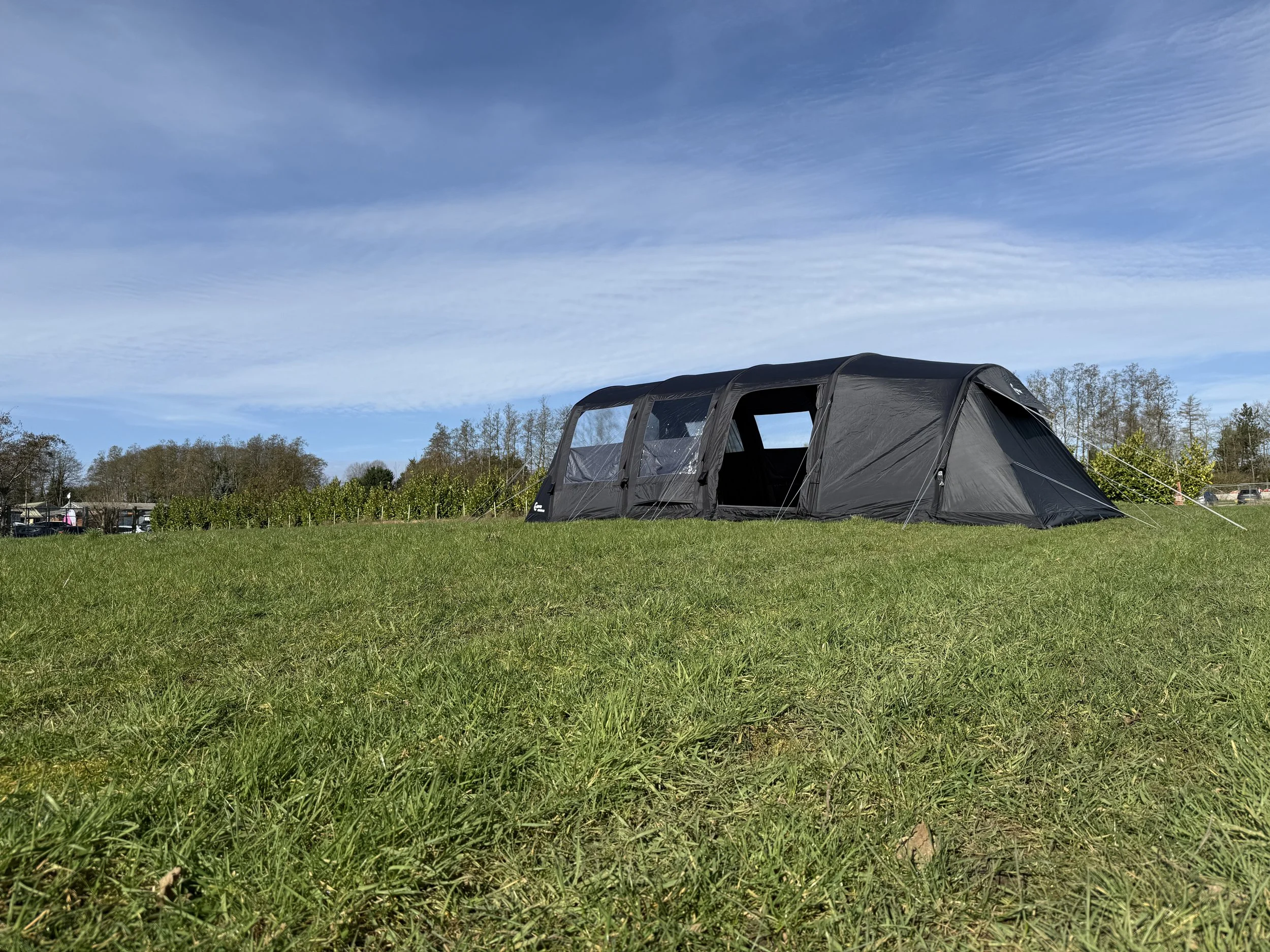 A black tent set up on green grass in a park or field on a clear day with a blue sky and some scattered clouds, surrounded by trees in the background.