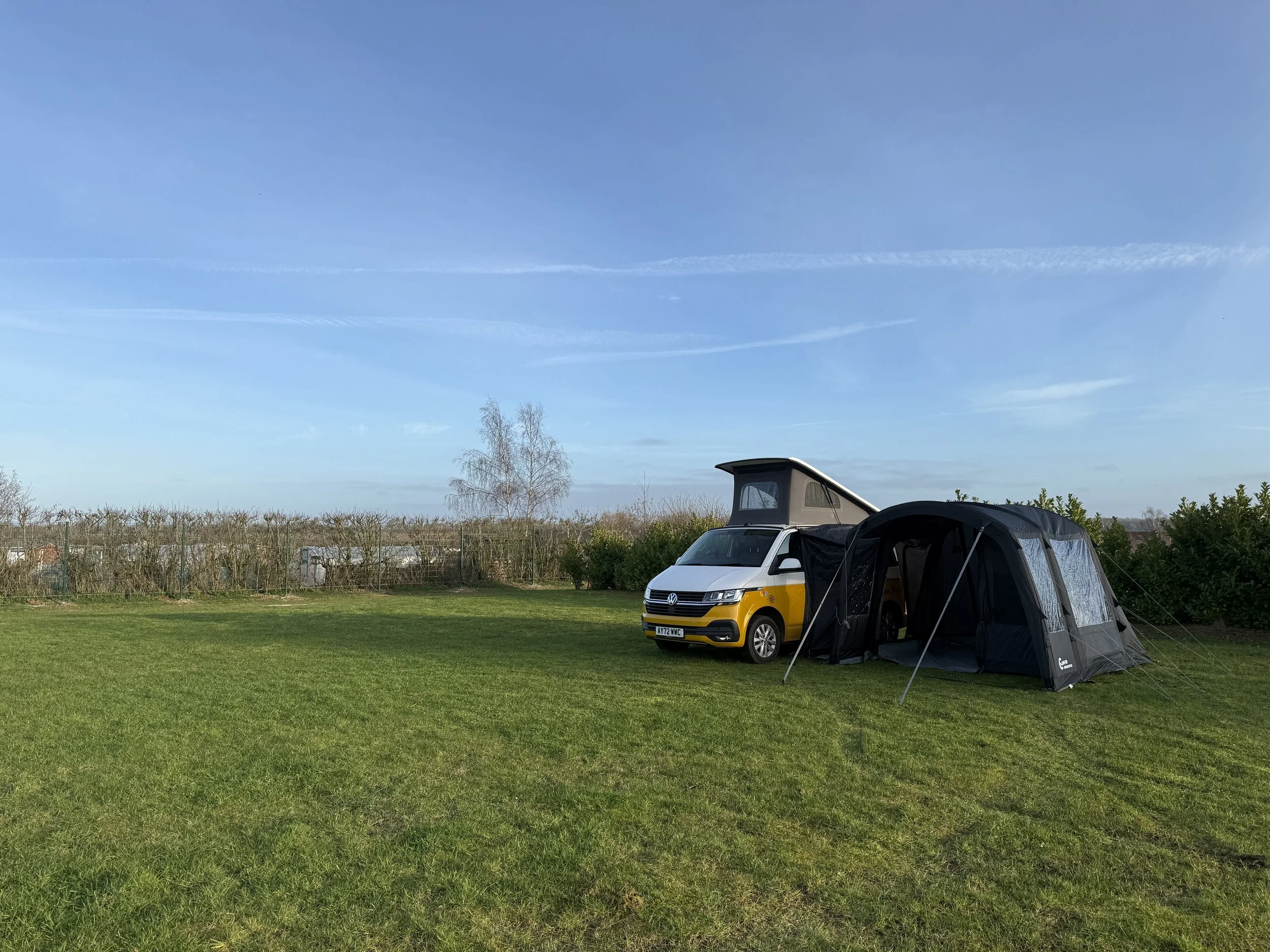 A camper van with a pop-up roof parked on a grassy field with a camping tent attached. In the background, there are trees, a fence, and a partly cloudy blue sky.