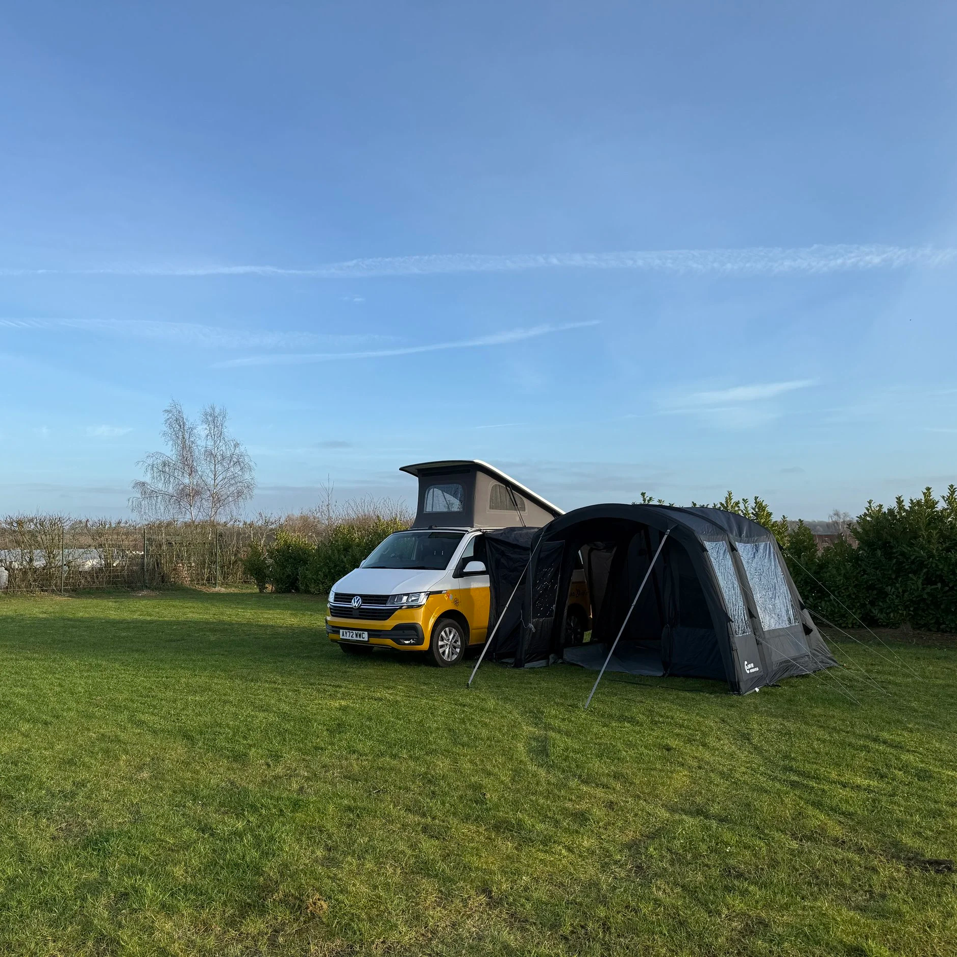 A yellow and white camper van parked on a grassy field with a tent set up next to it. In the background, there are trees and a blue sky with a few white clouds.