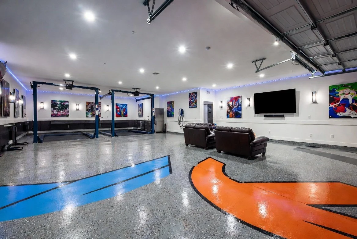 A spacious indoor recreational room with a concrete floor featuring painted orange and blue design lines, white walls decorated with colorful artwork, a large flat-screen TV, leather couches, a small bar area, and a mounted pair of racing harnesses. The room has bright ceiling lights, ambient wall lighting, and a garage-style door in the ceiling.