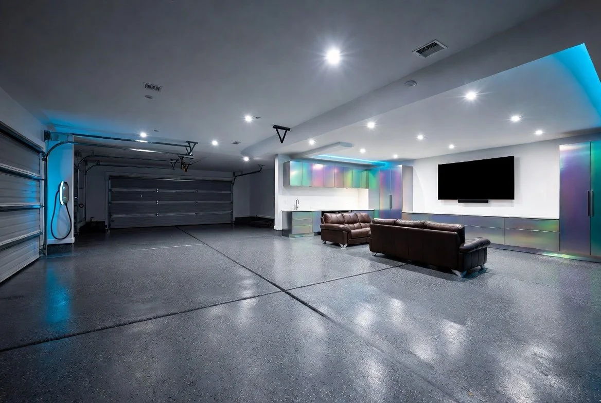 A spacious three-car garage with polished concrete floors, modern LED lighting, and metallic cabinets with iridescent finishes. There are two brown leather couches facing a large flat-screen TV mounted on the wall. The garage has a roll-up door and an electric charging station on the left side.