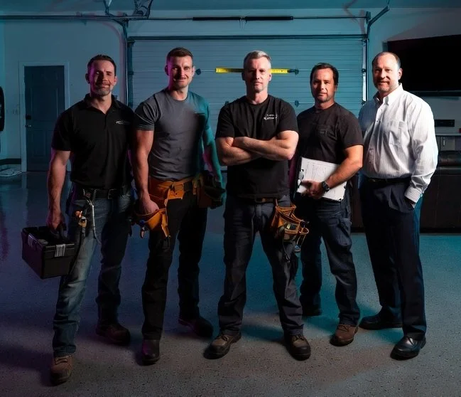 Five men standing in a garage, dressed in work clothes with tools, posing for a photo.