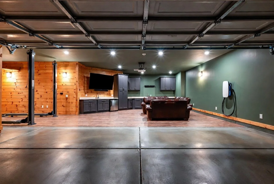 A spacious garage with a finished interior, including a section with wood paneling, a mounted flat-screen TV, gray cabinets, and a leather sofa. The garage door is open, revealing the concrete floor outside.