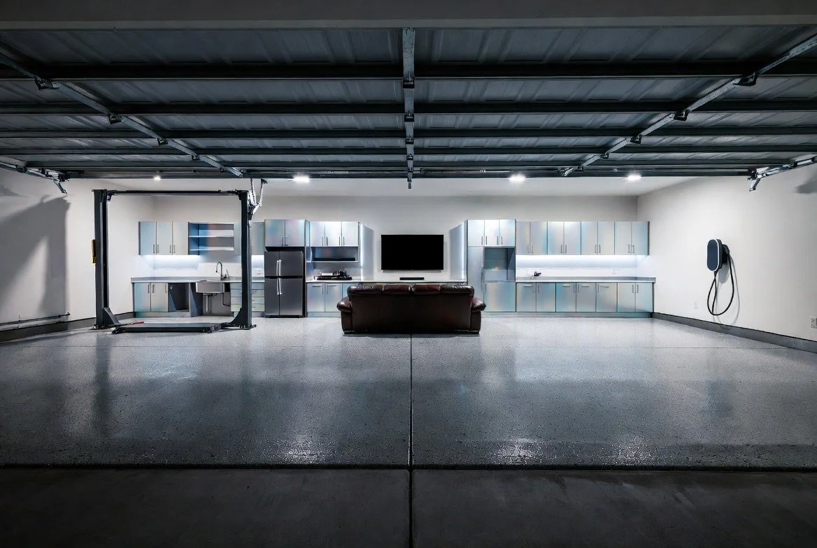 A spacious, clean garage featuring a mini kitchen with metallic cabinets, a refrigerator, a flat-screen TV, a leather couch, and a wall-mounted power washer.