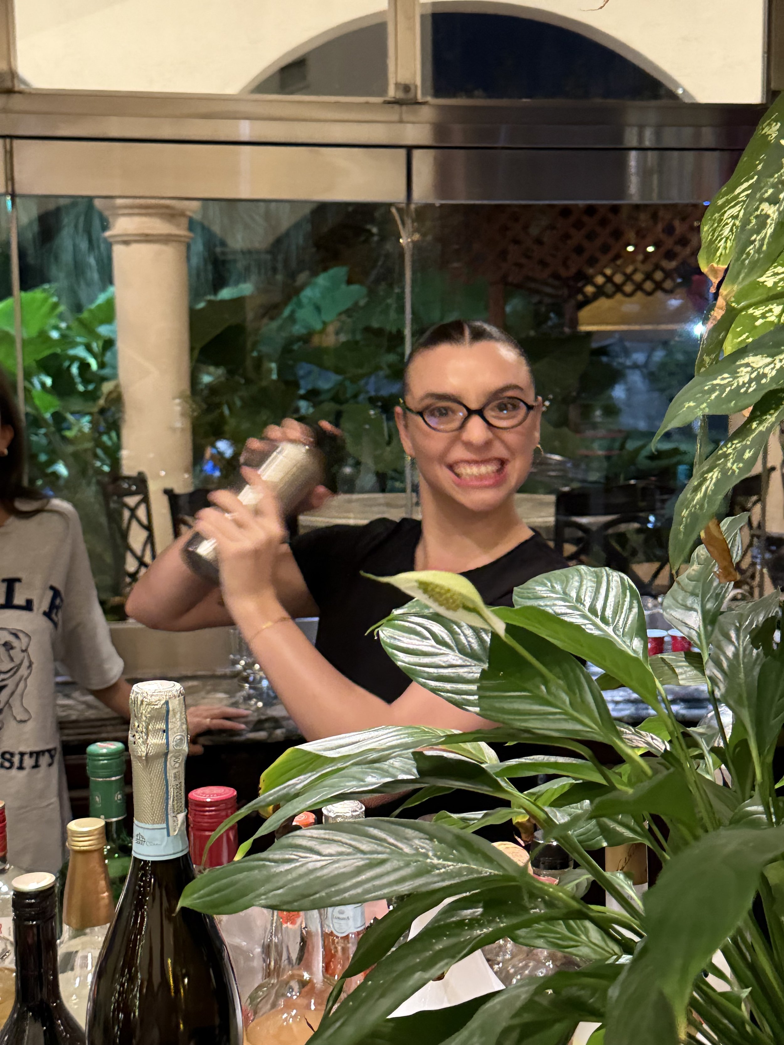 A woman with glasses and dark hair in a bun, wearing a black shirt, making a funny face while holding a cocktail shaker, inside a room with green plants and bottles on the counter.