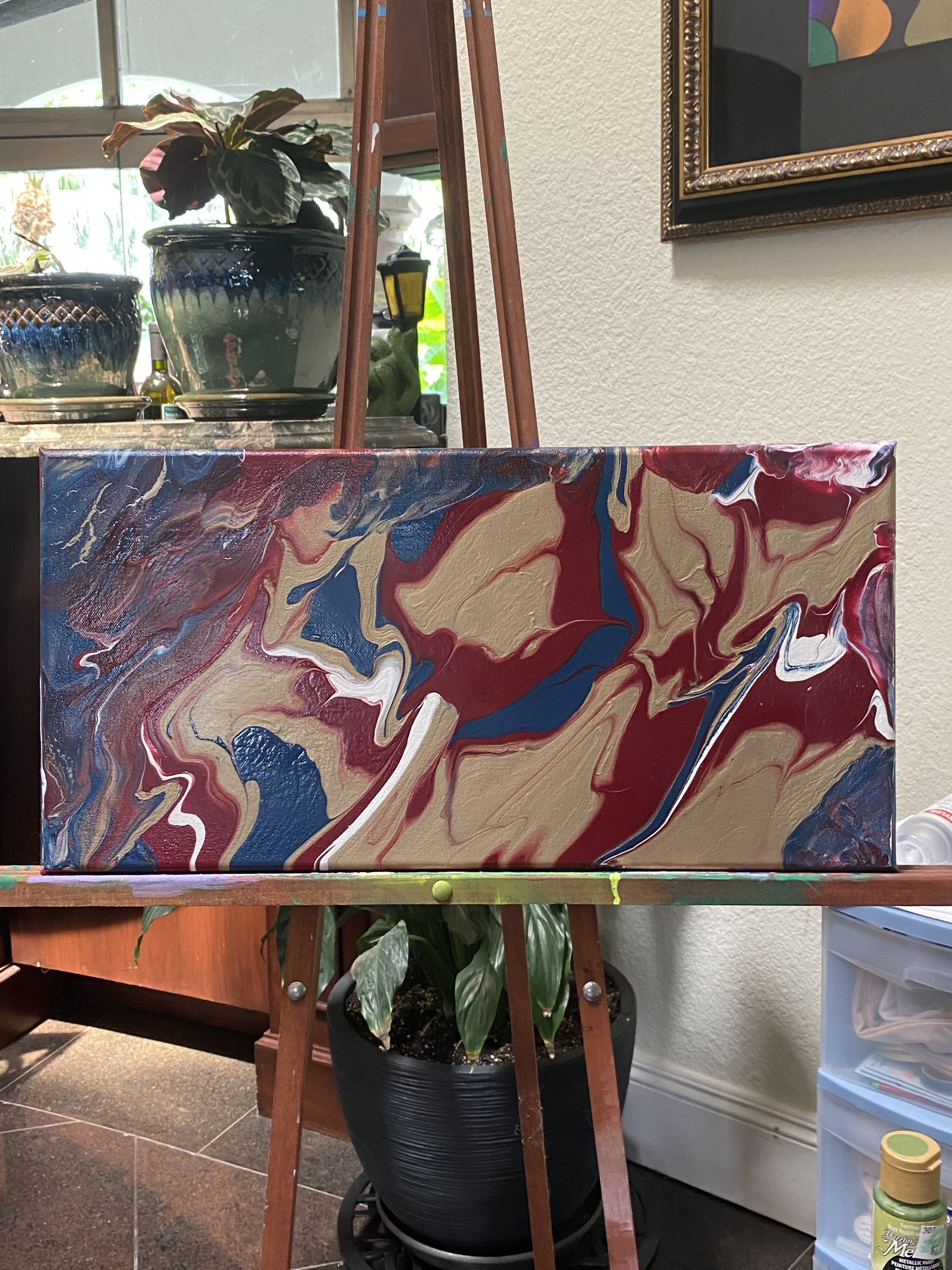 A rectangular canvas with an abstract swirl painting in red, beige, blue, white, and black, placed on a wooden easel in a room with potted plants and framed artwork on the wall.