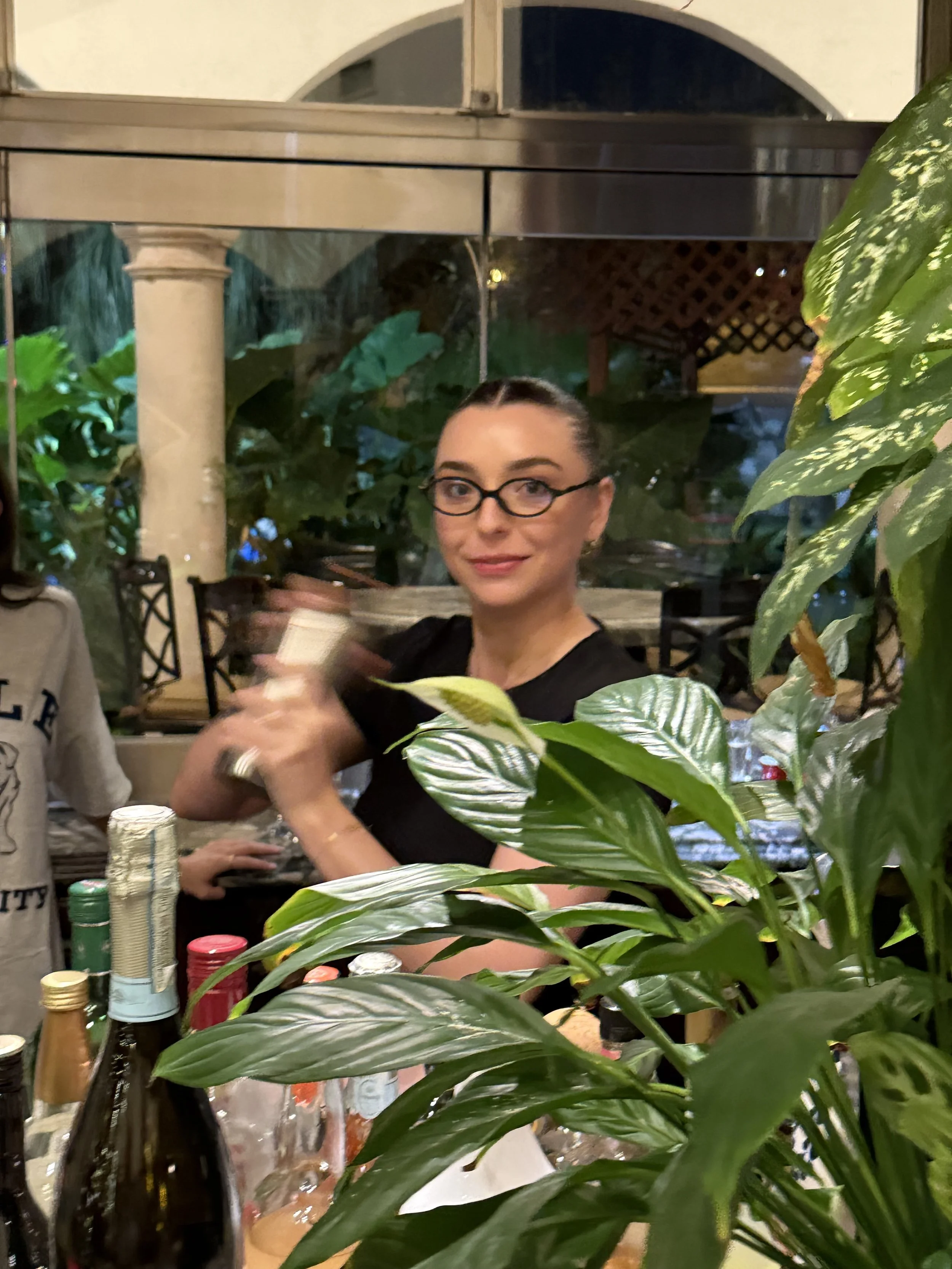 A woman with glasses and dark hair tied back, sitting at a table with green plants and bottles, looking at the camera inside a restaurant or cafe at night.