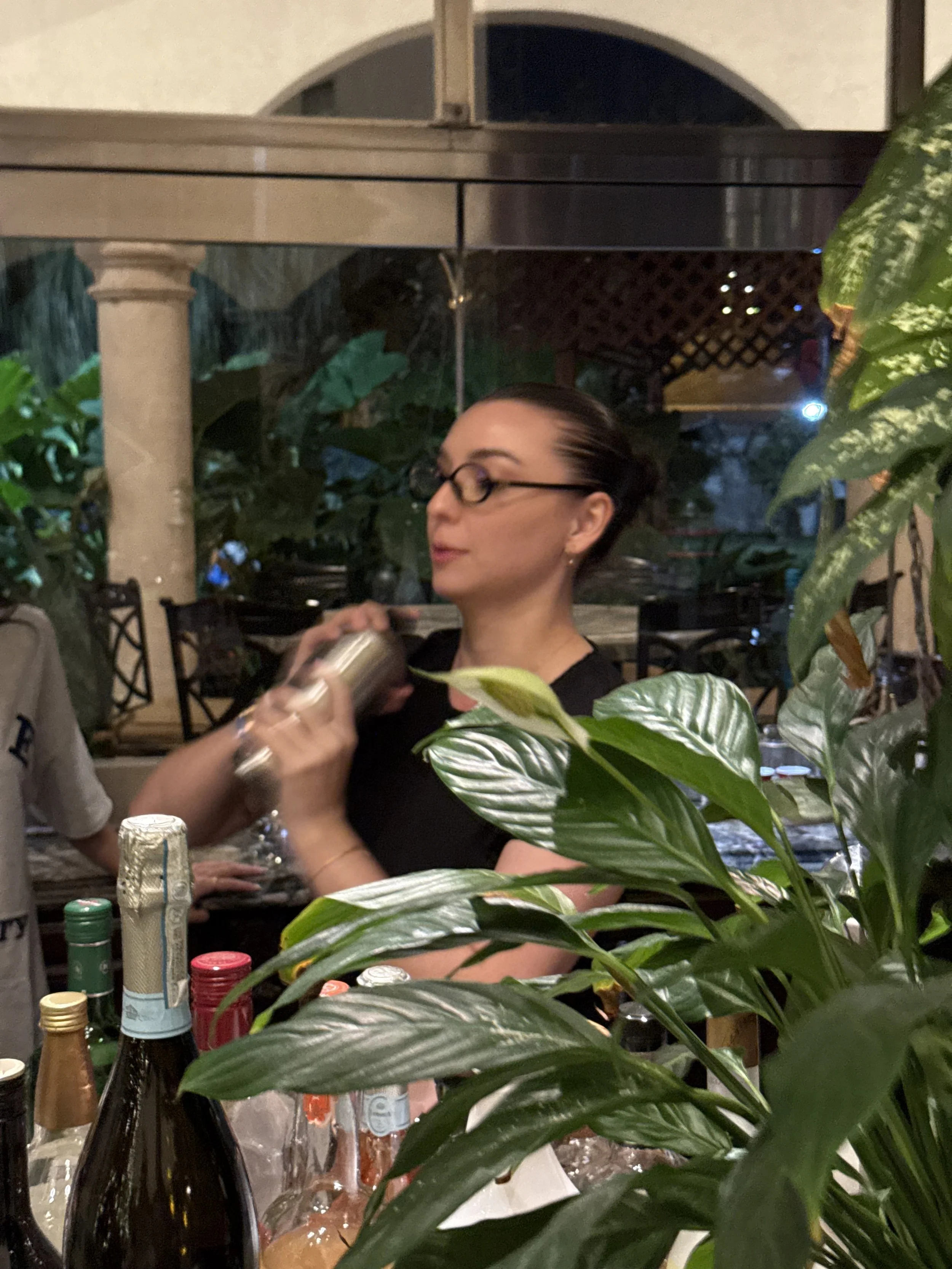 A woman with glasses and dark hair tied back, standing behind a bar with various bottles, surrounded by green plants, as she shakes a cocktail shaker. The setting appears to be a restaurant or bar at night.