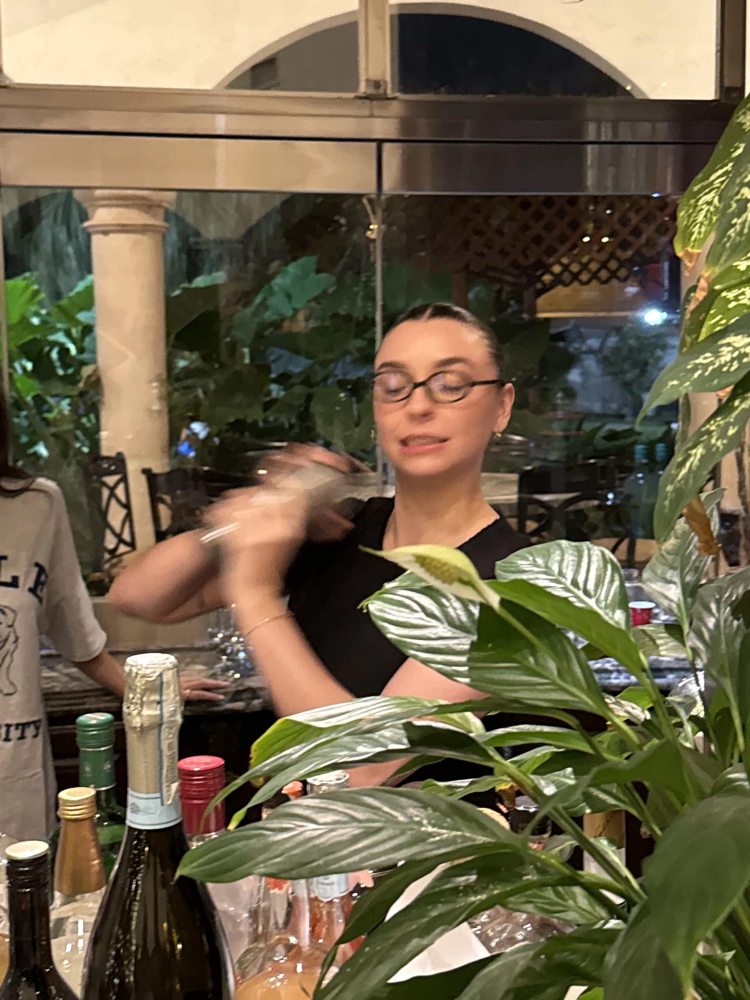 A woman with glasses and dark hair pulled back, wearing a black shirt, standing behind a bar with bottles and plants in the foreground, in an indoor setting with large windows and greenery outside.