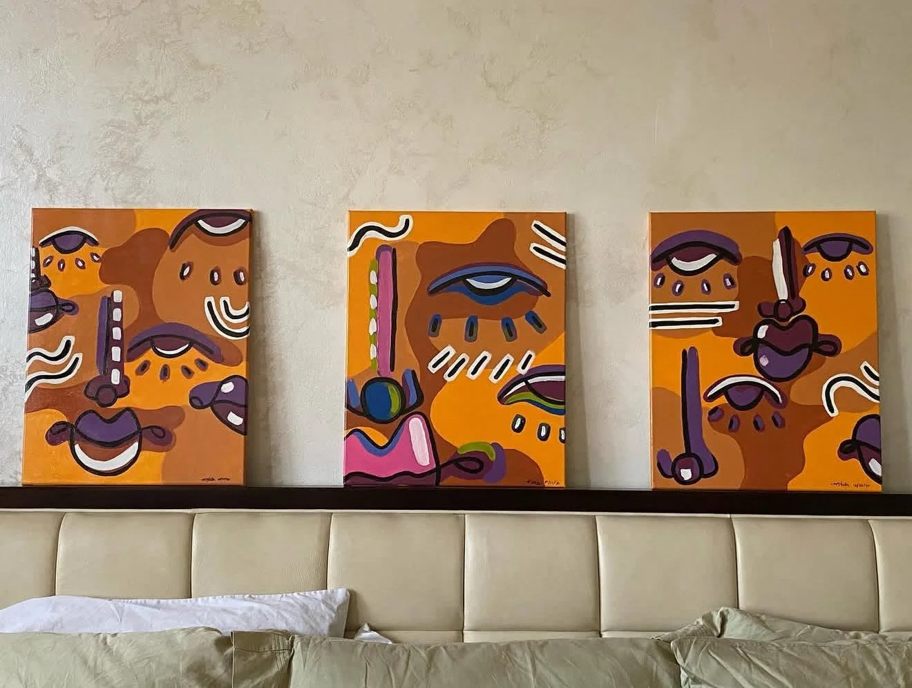 Three abstract paintings with orange backgrounds and bold black, white, and purple lines, hanging above a bed with beige pillows and headboard.