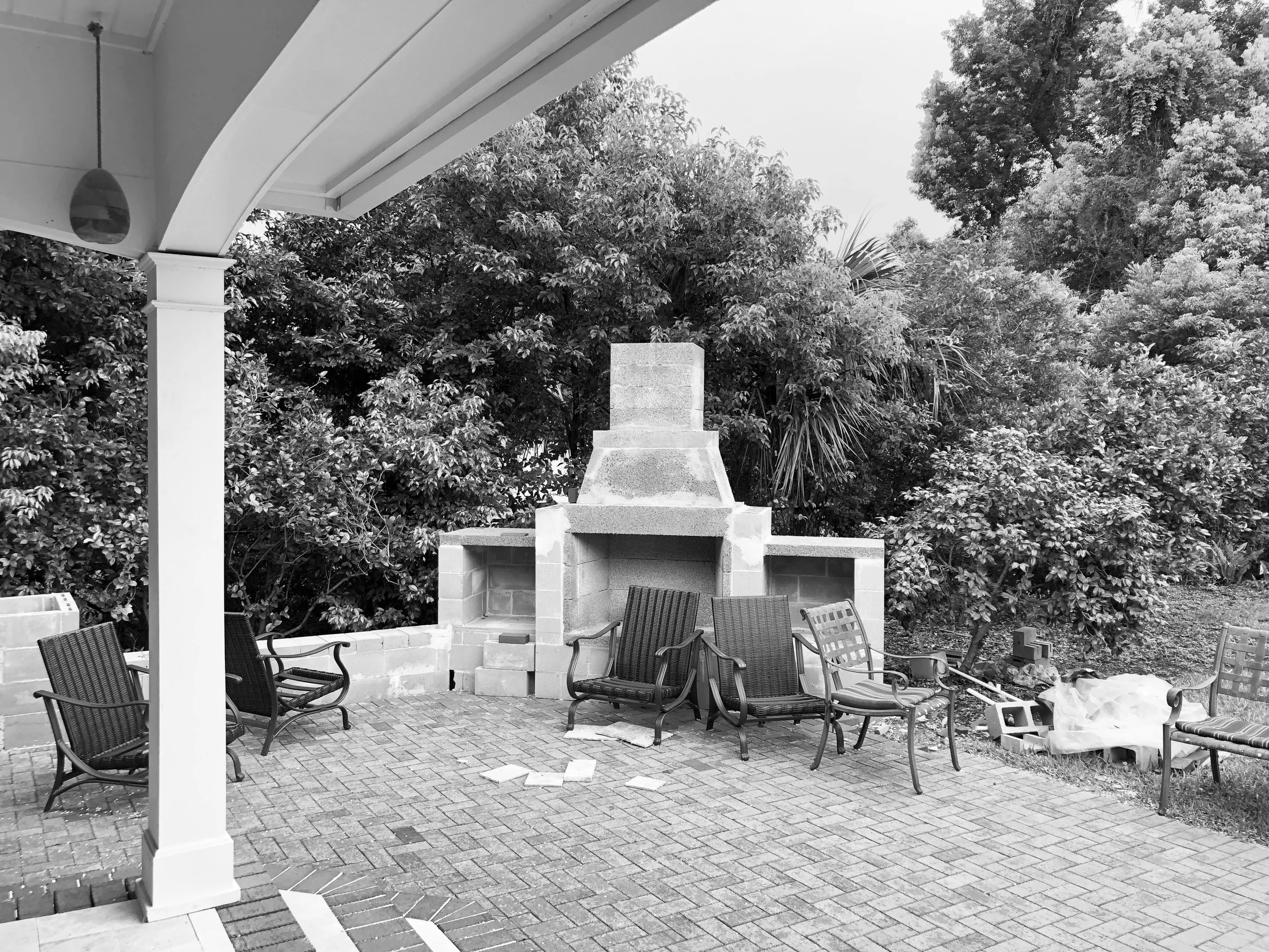 Black and white photo of a backyard patio with several chairs arranged around a brick-paved area. A stone outdoor fireplace and lush trees in the background. Some bricks and other materials are placed on the ground near the chairs, indicating ongoing