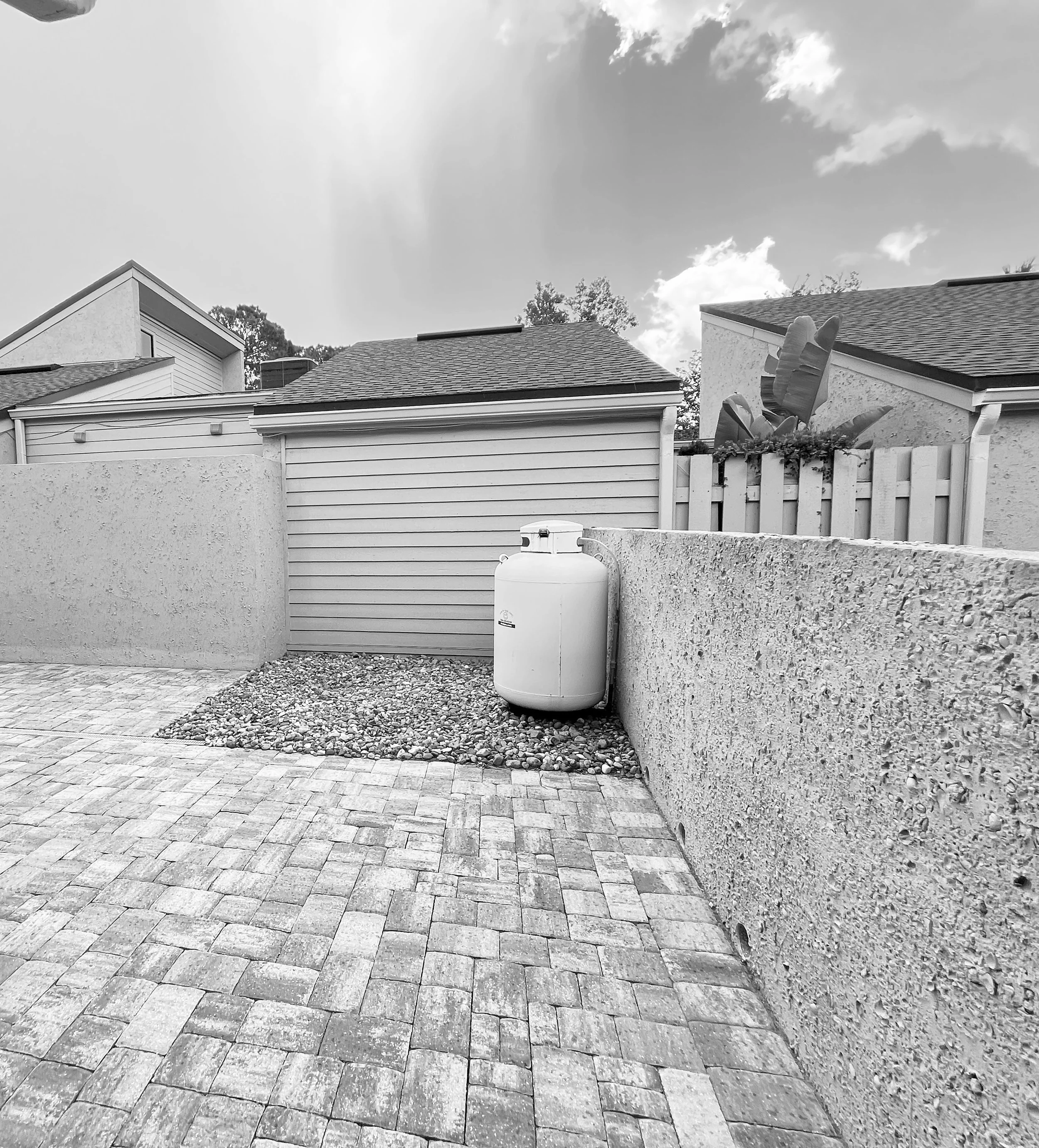 A backyard with a brick patio, concrete wall, small shed with a roll-up door, a propane tank, and some trees and plants behind a white picket fence.