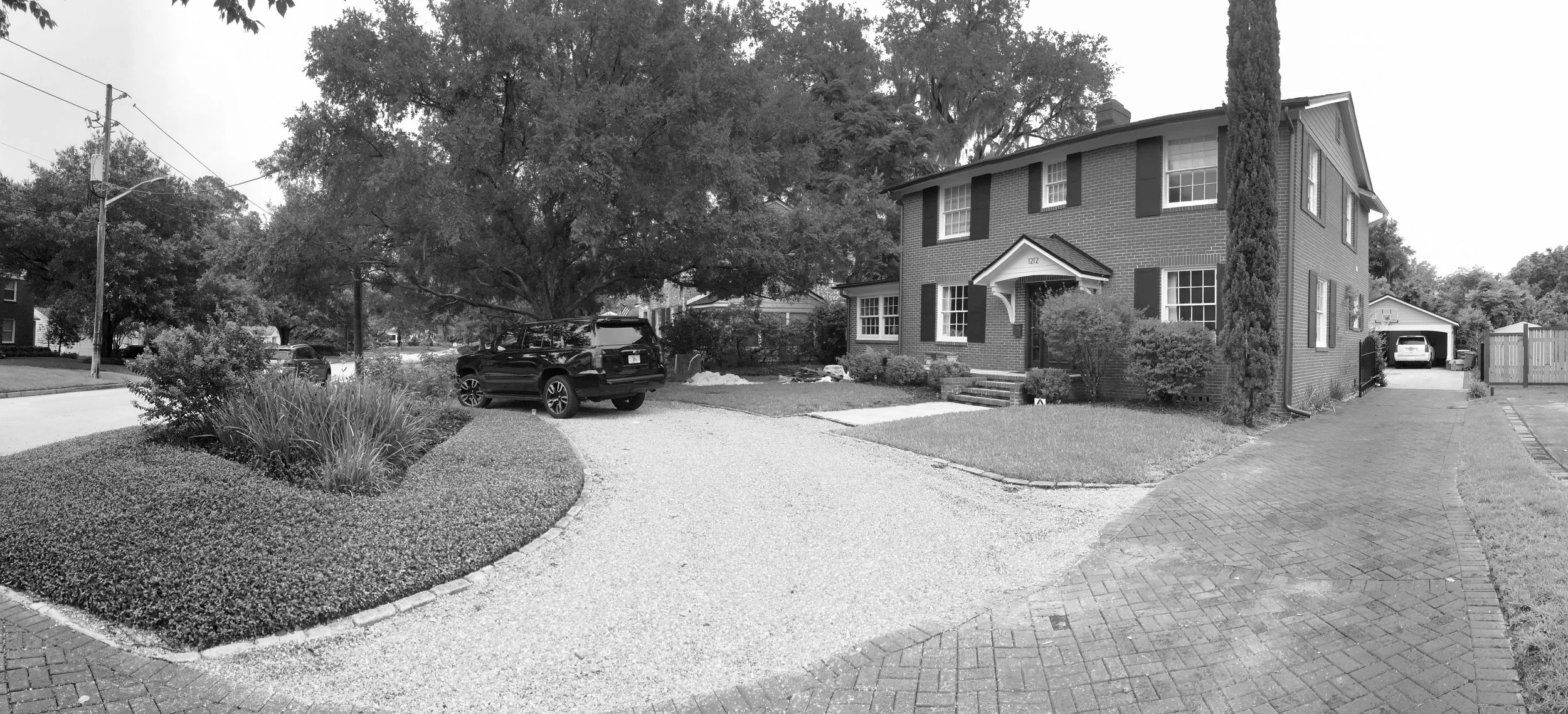A two-story brick house with a front porch and a driveway with parked cars. There are bushes, grass, and tall trees in the yard, along with a paved sidewalk and a neighboring house visible in the background.