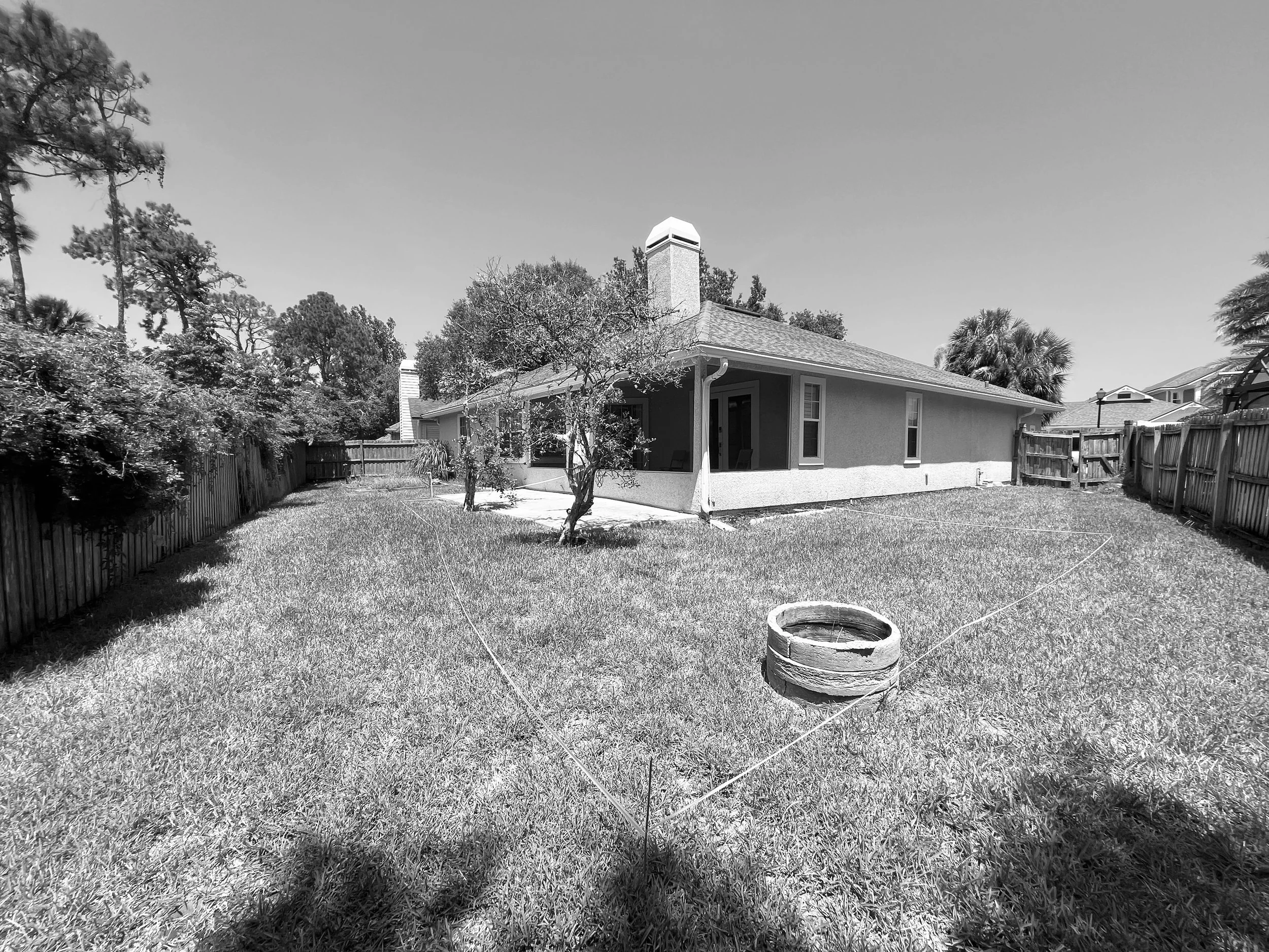 Backyard with grass, trees, wooden fence, and a house with a chimney, seen in black and white.