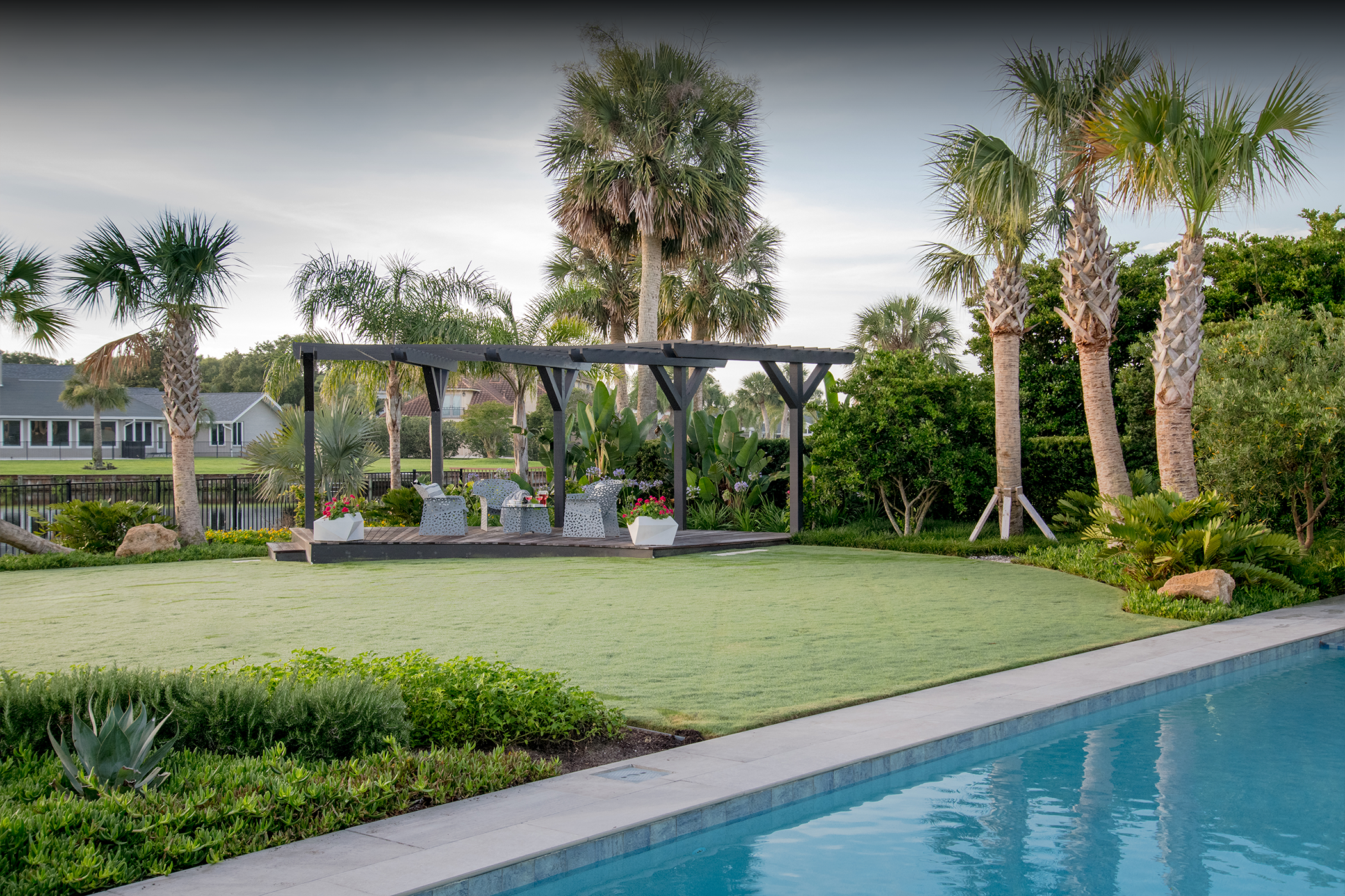 A backyard with a swimming pool, a well-maintained lawn, and a patio with outdoor furniture under a pergola, surrounded by various palm trees and lush greenery.