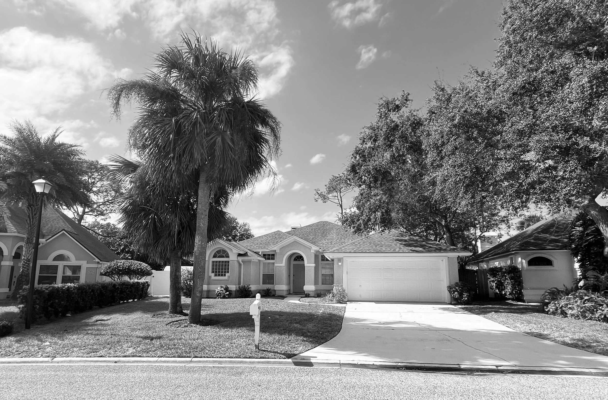 A residential house with a driveway, garage, and trees in the front yard, including a palm tree and leafy trees, under a partly cloudy sky.